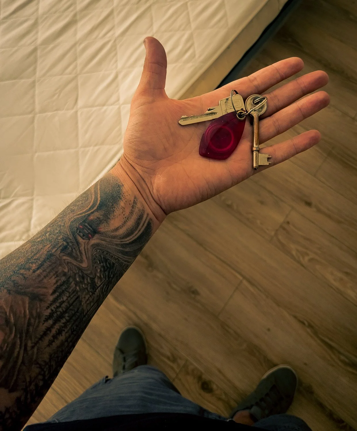 A person with a tattooed arm holding three keys, including a house key, a skeleton key, and a red keychain key, all on a keyring.