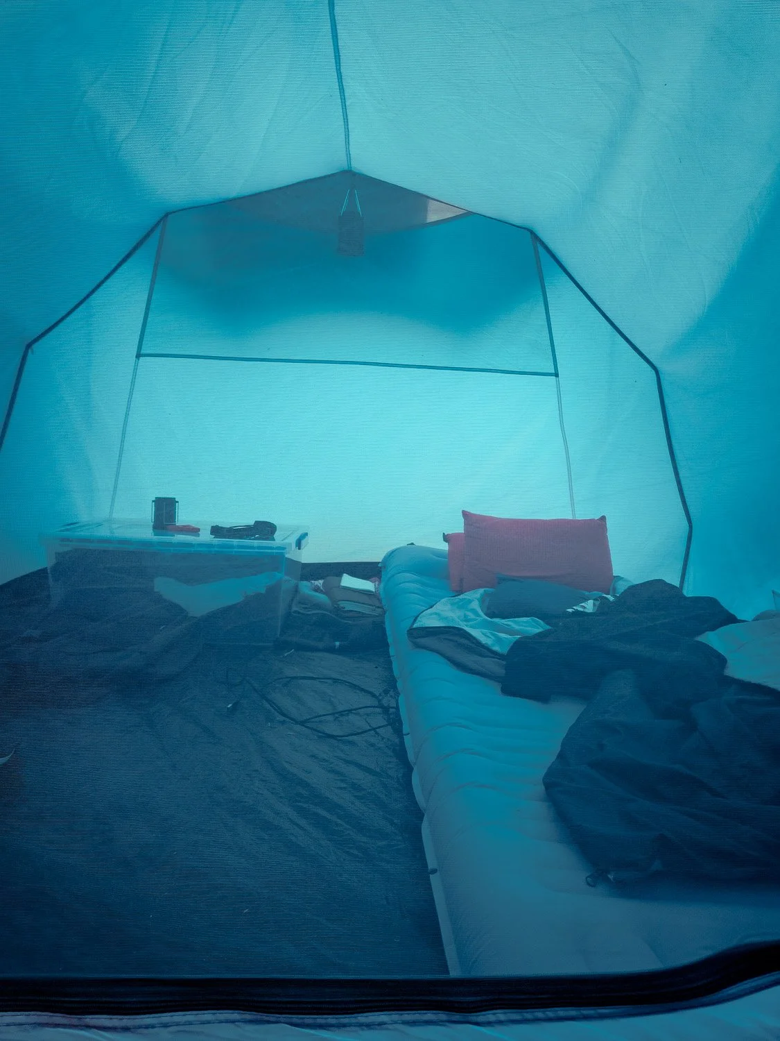 Interior of a tent with a bed, pillows, and scattered clothes, viewed through a mesh window.