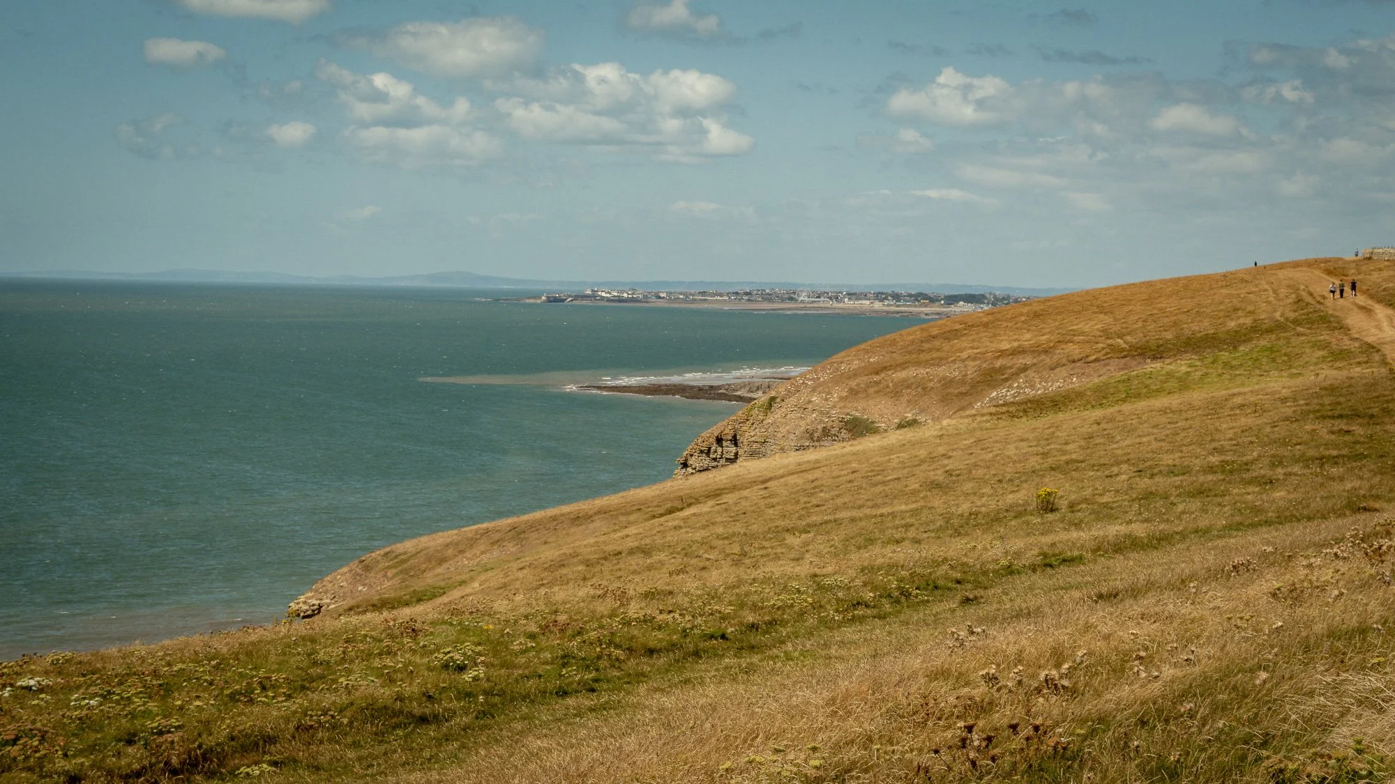 A coastal cliff with grassy terrain and a walking trail with a few people, overlooking the ocean and a distant town on the horizon, under a partly cloudy sky.