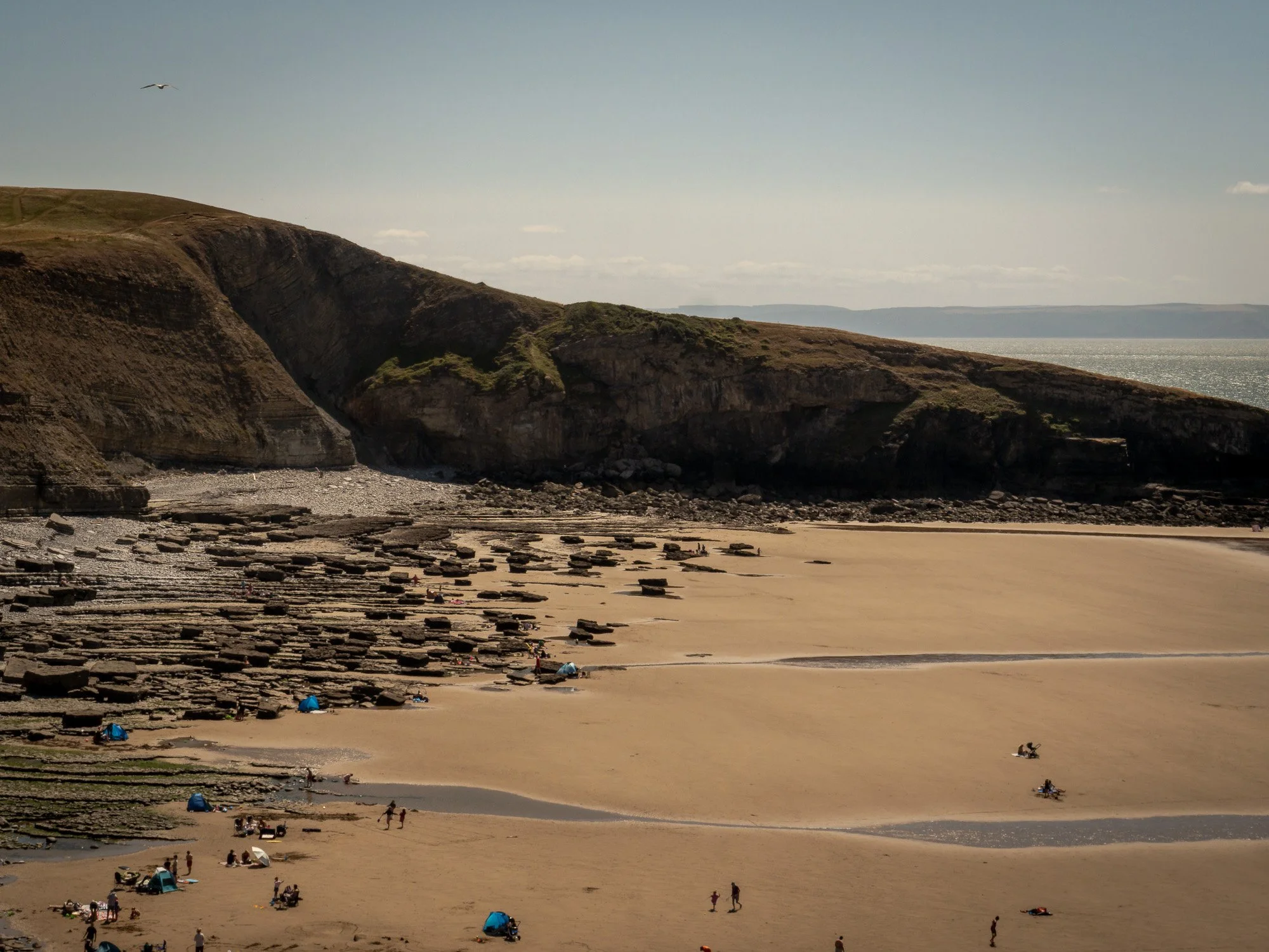 A coastal scene with sandy beach, people, tents, rocky formations, green cliffs, and ocean with a cloudy sky.