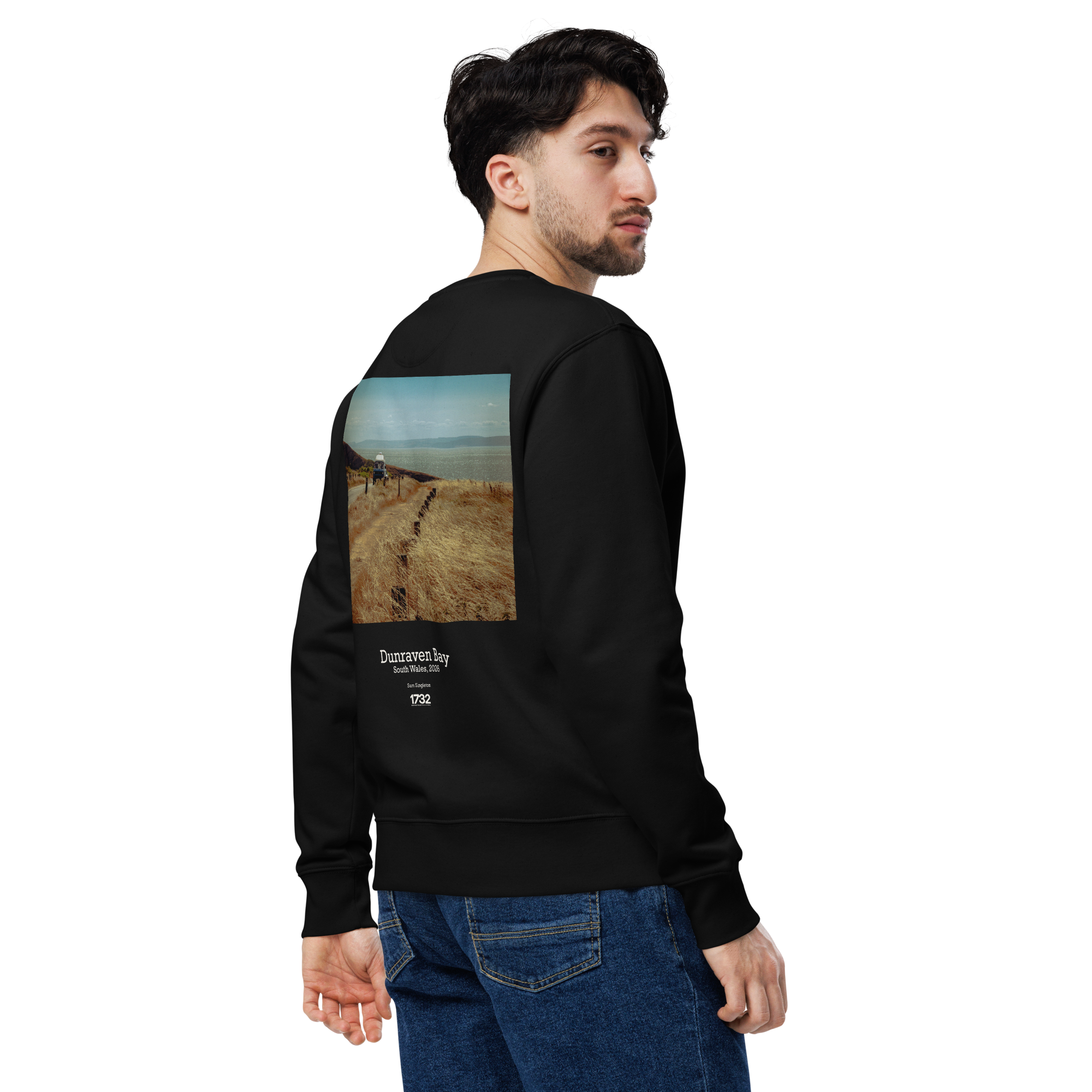 A young man with dark hair and a beard wearing a black sweatshirt with landscape and text art on the back, standing in front of a black backdrop.