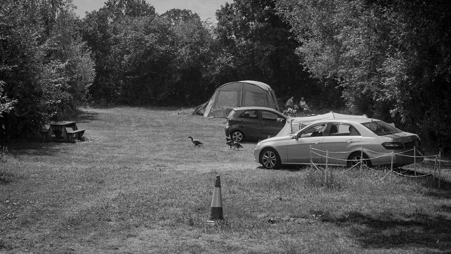 Camping area with three parked cars, a tent, a picnic table, a traffic cone, and two people sitting on the ground next to trees in the background.
