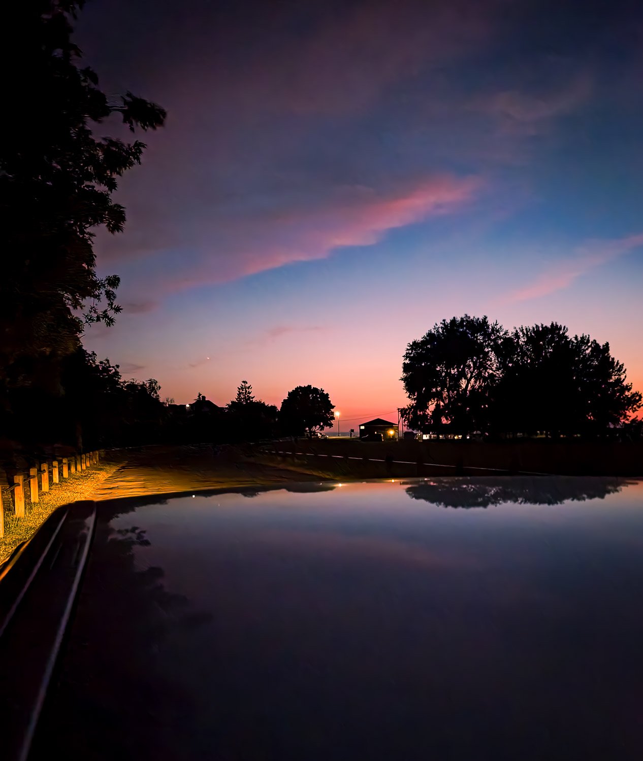 Sunset or sunrise over a rural scene with trees, a road, and a few buildings, reflected on the roof of a vehicle in the foreground.
