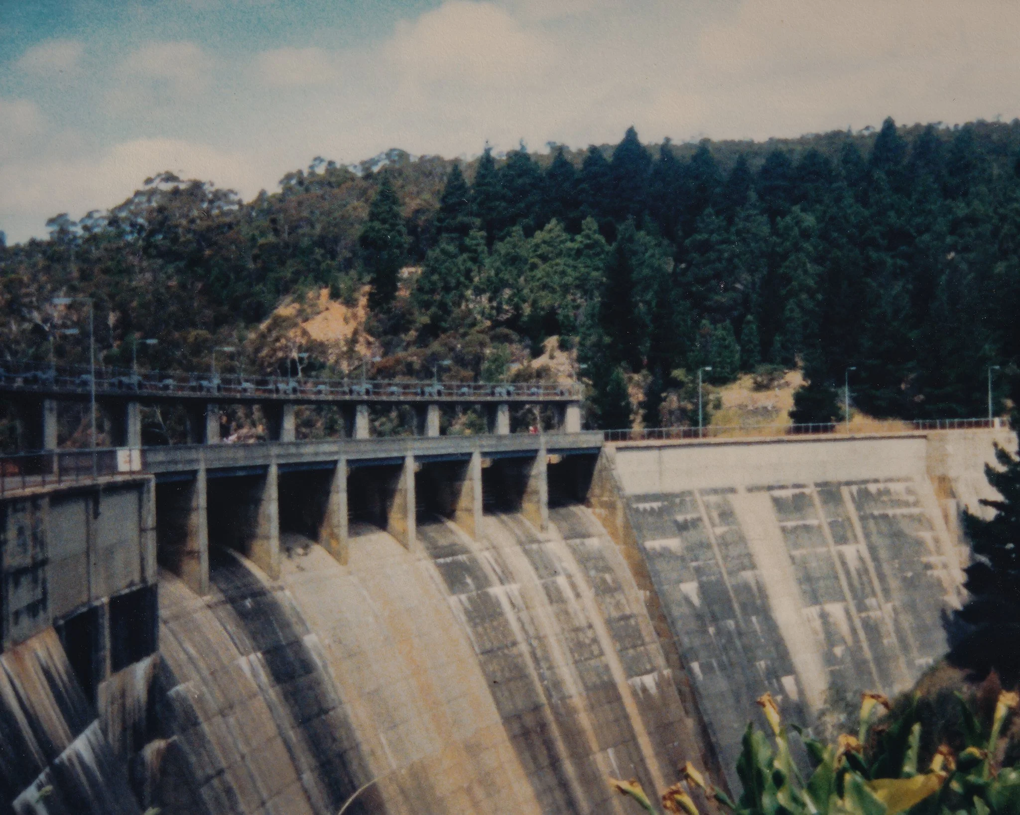 A large dam with concrete spillways and a highway bridge running across it, with green forested hills in the background.