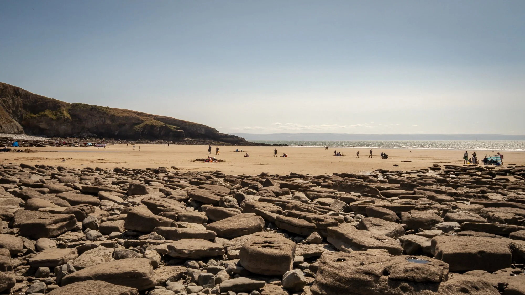 A sandy beach with large rocks in the foreground, people relaxing and walking, cliffs on the left, and the ocean under a clear sky in the background.