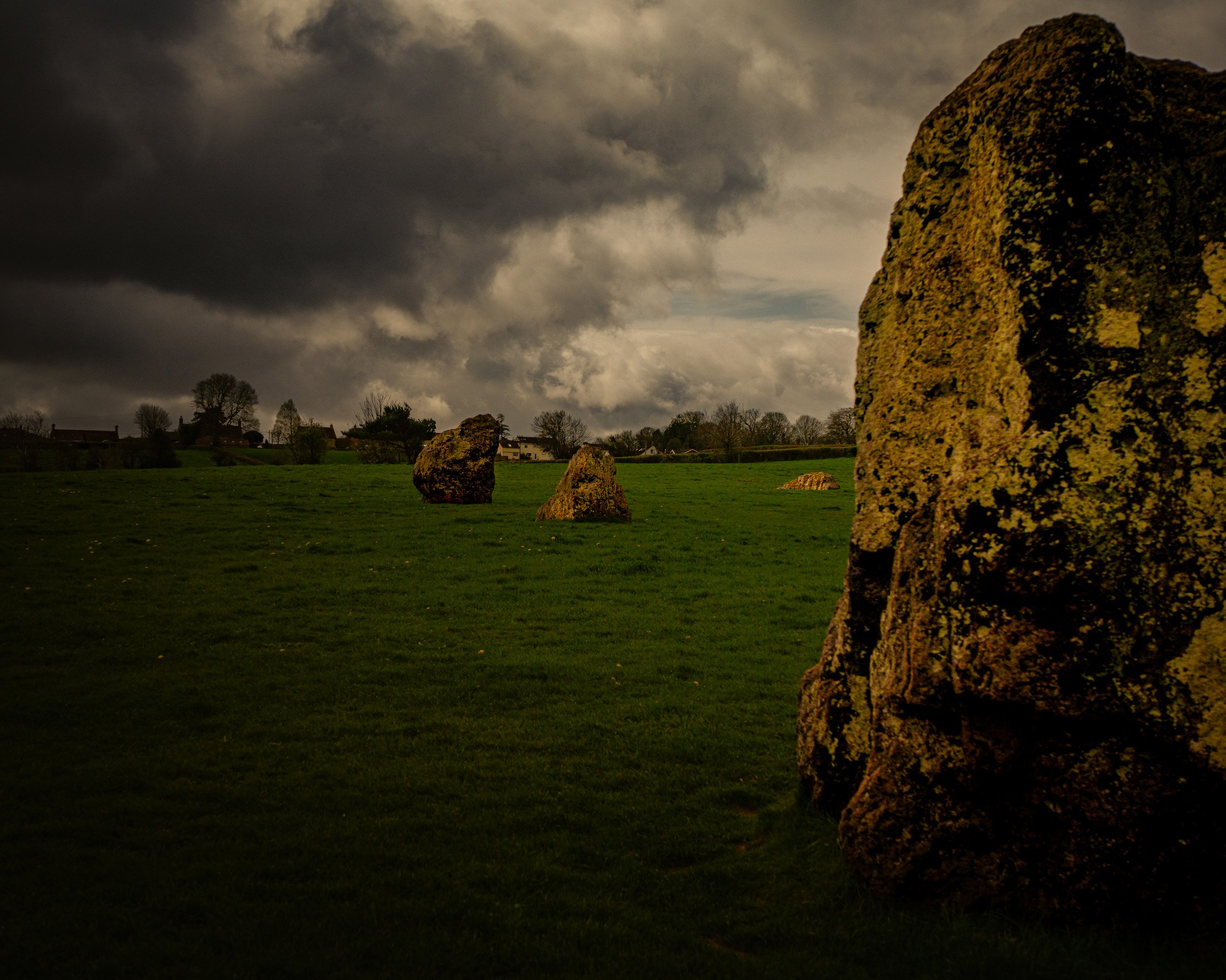 Large moss-covered standing stone in a grassy field with smaller stones nearby. Dark stormy sky with thick clouds overhead.