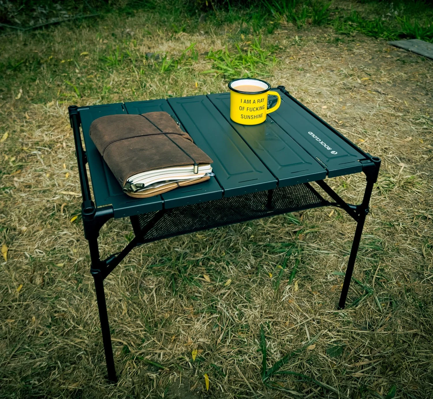 A black portable table on grassy ground. On the table, there is a brown rolled-up towel, a folded notebook or journal, and a yellow mug with black text that reads, 'I AM A RAY OF F***ING SUNSHINE'.