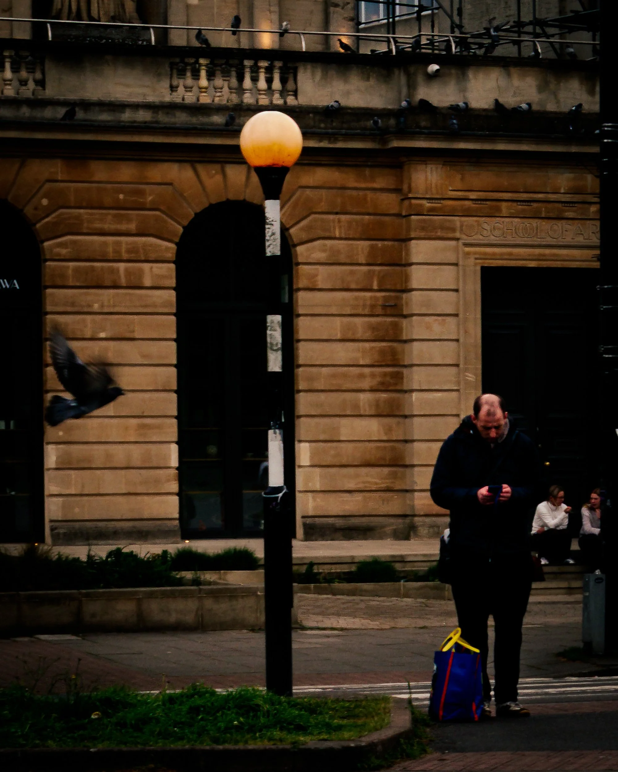 A man standing outdoors on a city sidewalk at dusk, looking at his phone, next to a blue and yellow bag, with a shopping cart and a group of people sitting on the ground nearby, in front of a large stone building with a lit doorway and a lamp post.