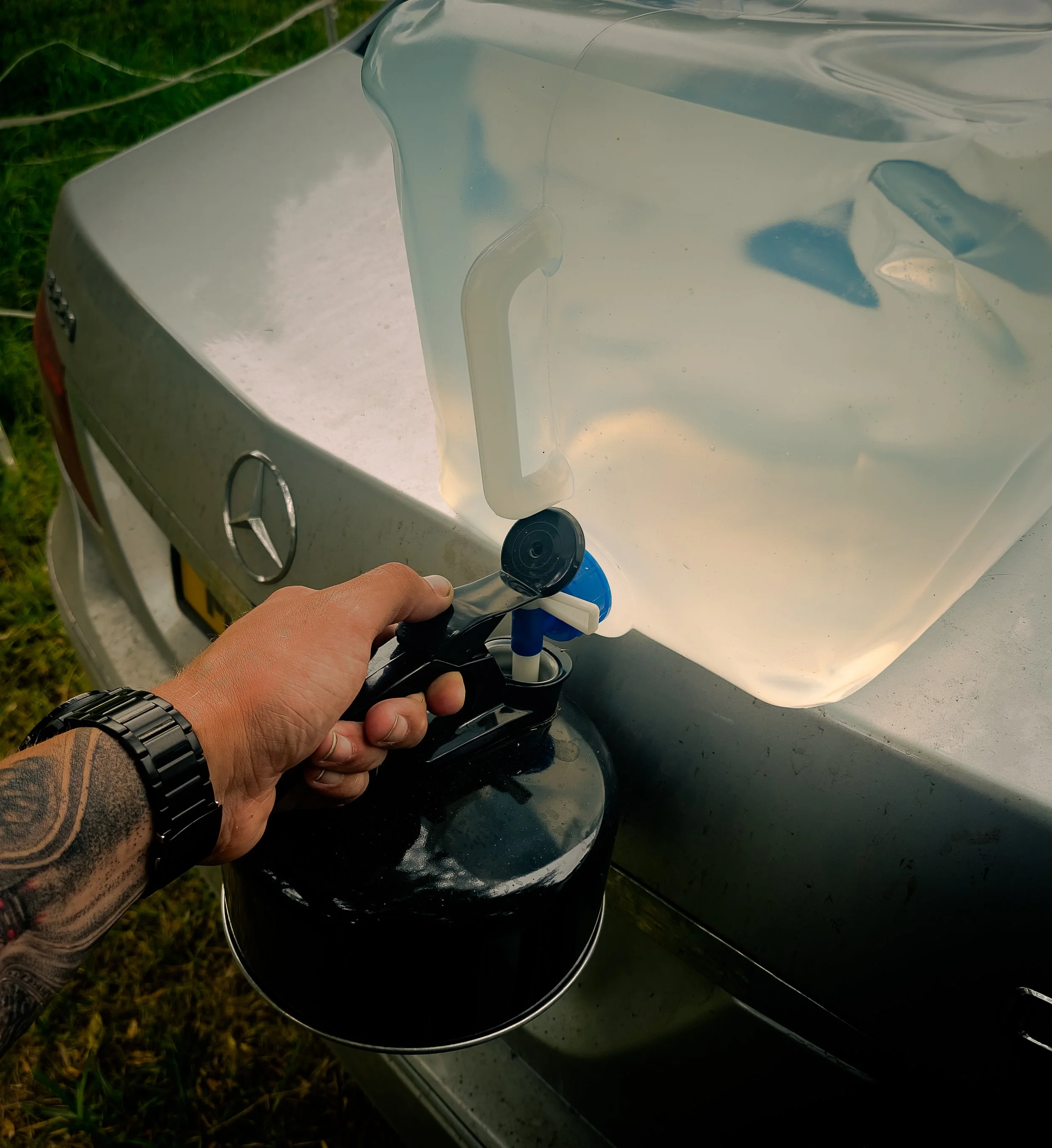A person with tattoos on their arm holding a device over a reflective, black motorcycle fuel tank, outdoors on grass and concrete.