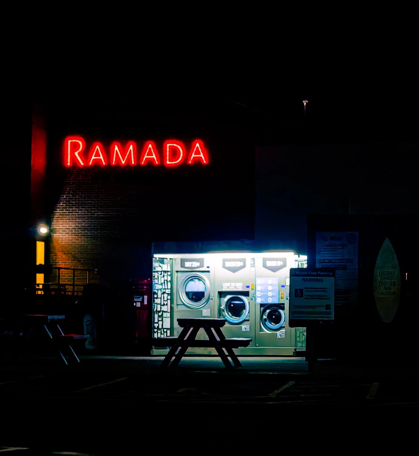 Nighttime view of a laundromat with bright washers inside, illuminated beneath a lit 'Ramada' neon sign.