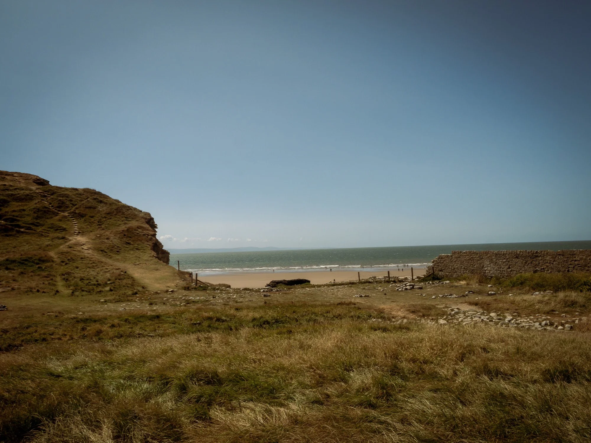 A scenic coastal landscape with grassy hills in the foreground, a sandy beach, and the ocean in the background under a clear blue sky.