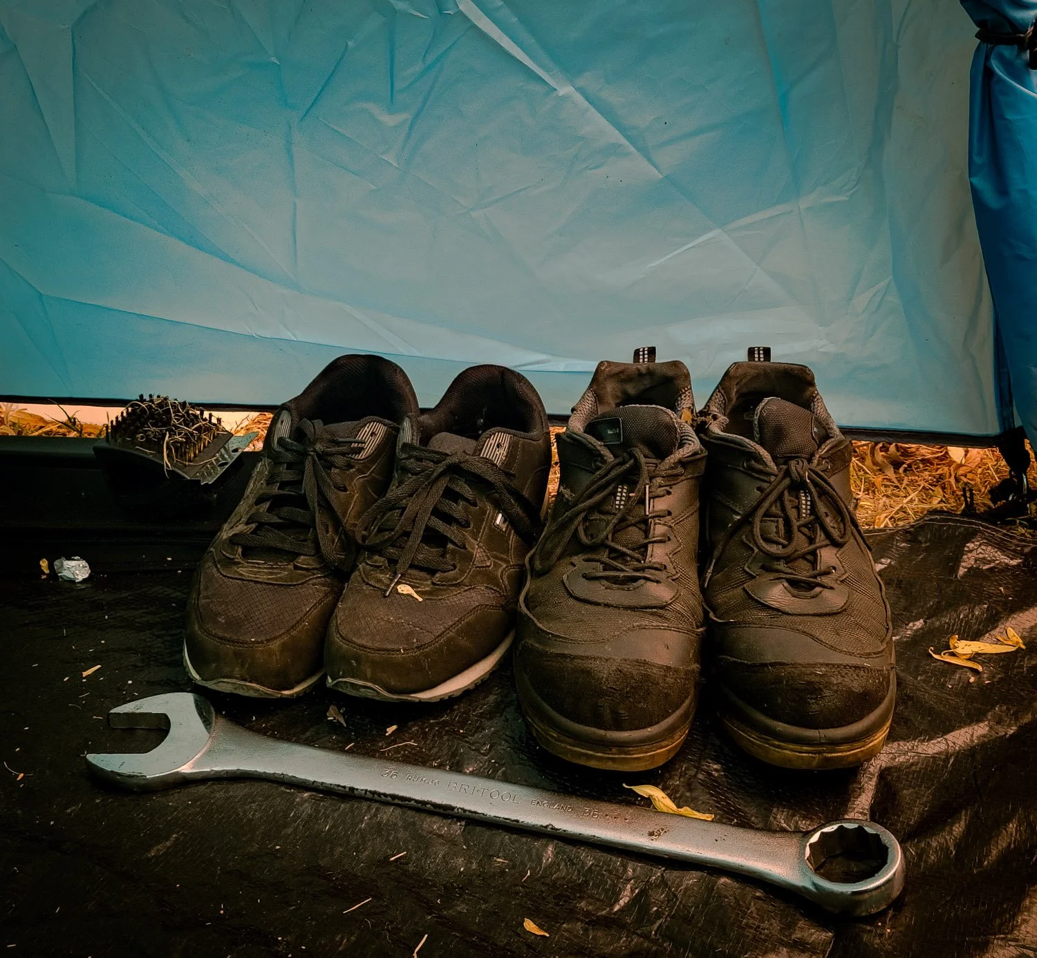 Pair of muddy brown hiking shoes on a black surface, with a wrench nearby and a blue tent in the background.