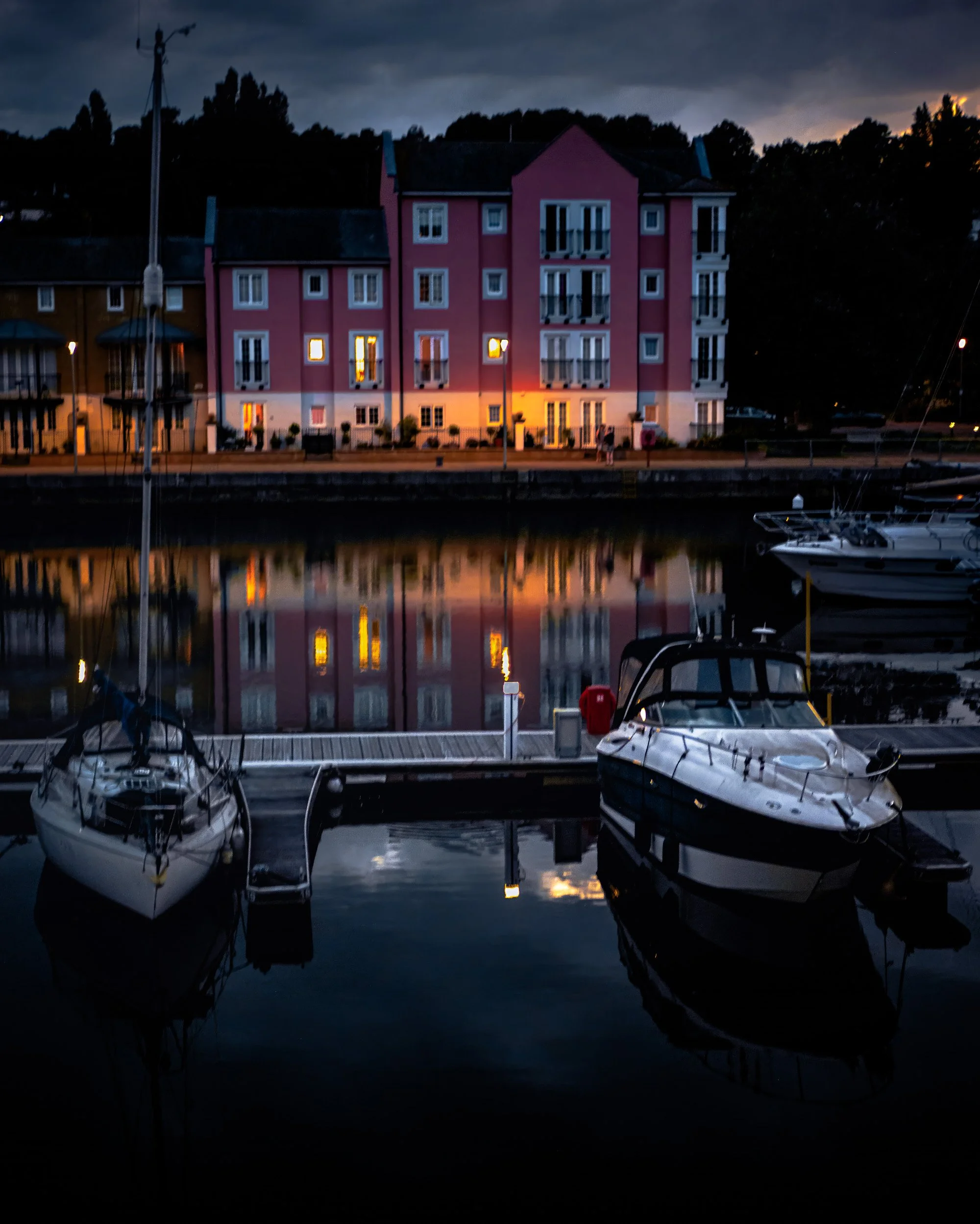 Night scene of a pink residential building by a water marina with boats, dark stormy sky in the background, and reflections of the building's lights on the water.