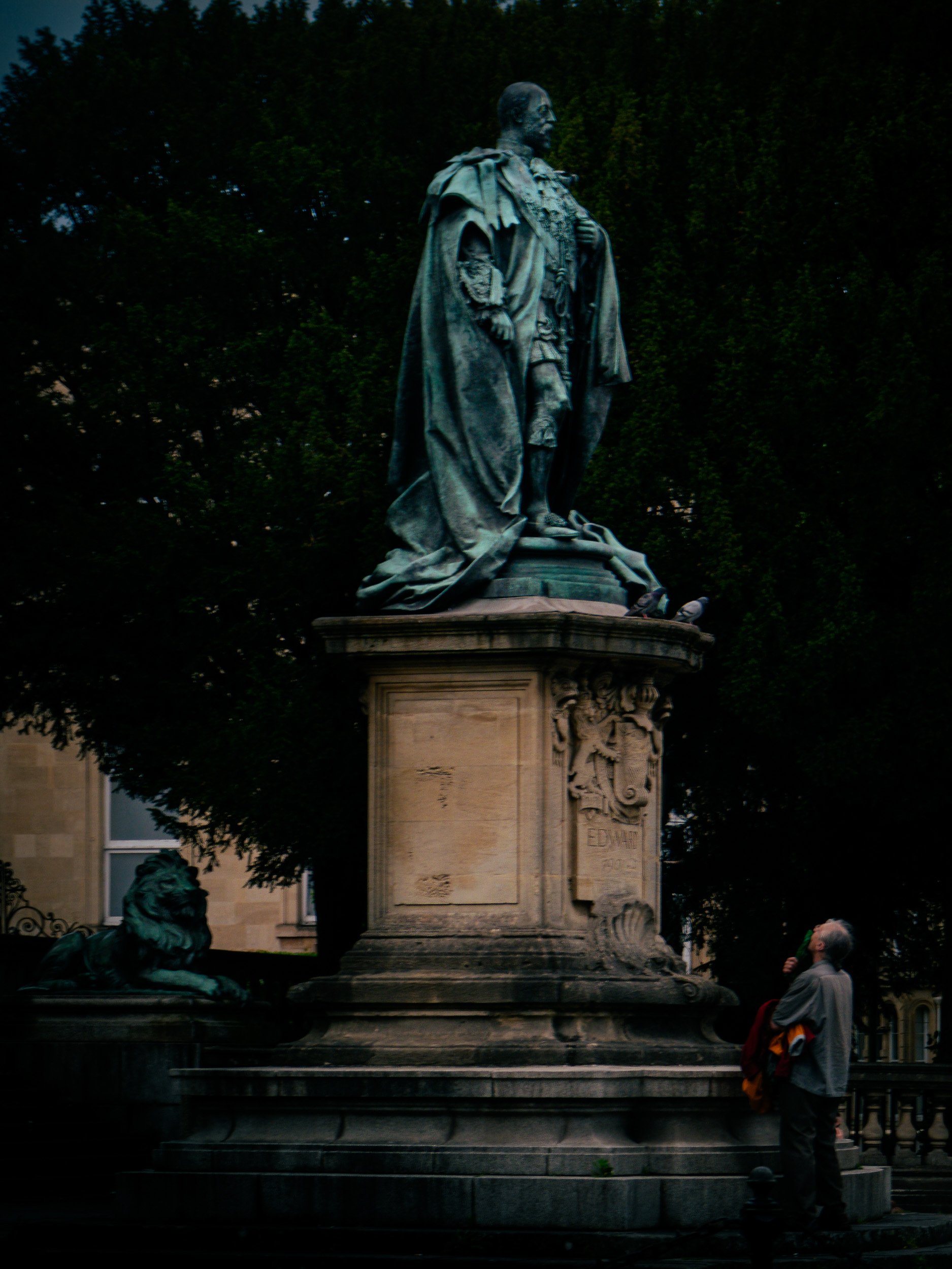 A statue of a man in military uniform with a cape, standing on a pedestal adorned with inscriptions and coats of arms. There is a dog sculpture at the base of the pedestal, and a person is standing nearby looking at the statue. The background feature