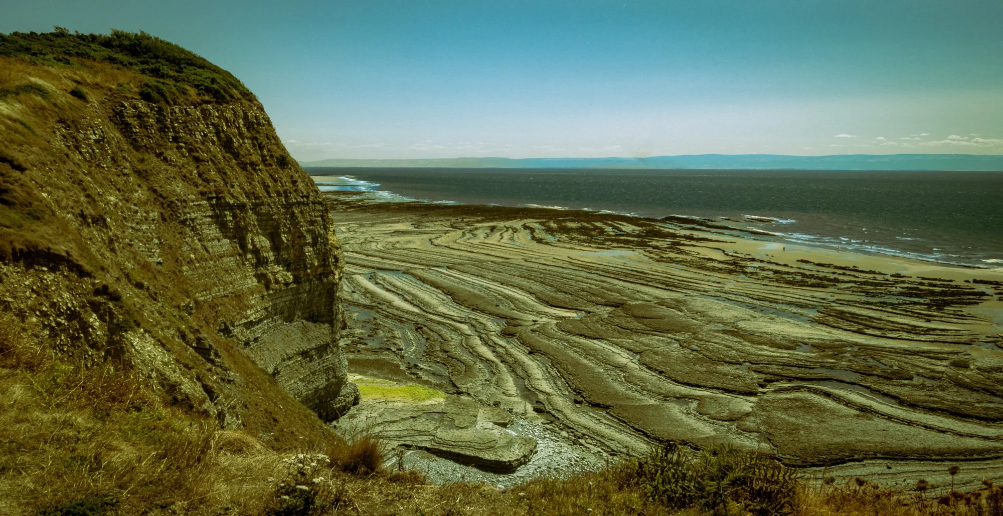 Coastal scene with steep cliff on the left, layered rock formations on the sandy shore below, and the ocean extending to the horizon under a clear blue sky.
