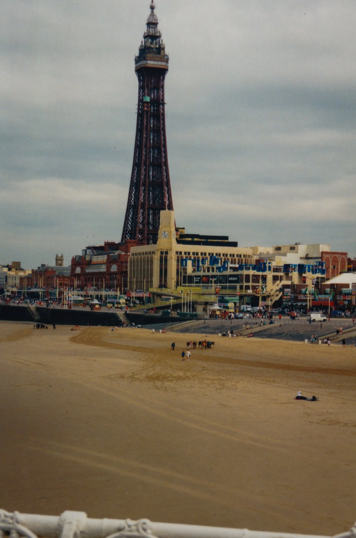 The Blackpool Tower in Blackpool, England, with a sandy beach and people walking or relaxing nearby, and a nearby building with a clock and shops.