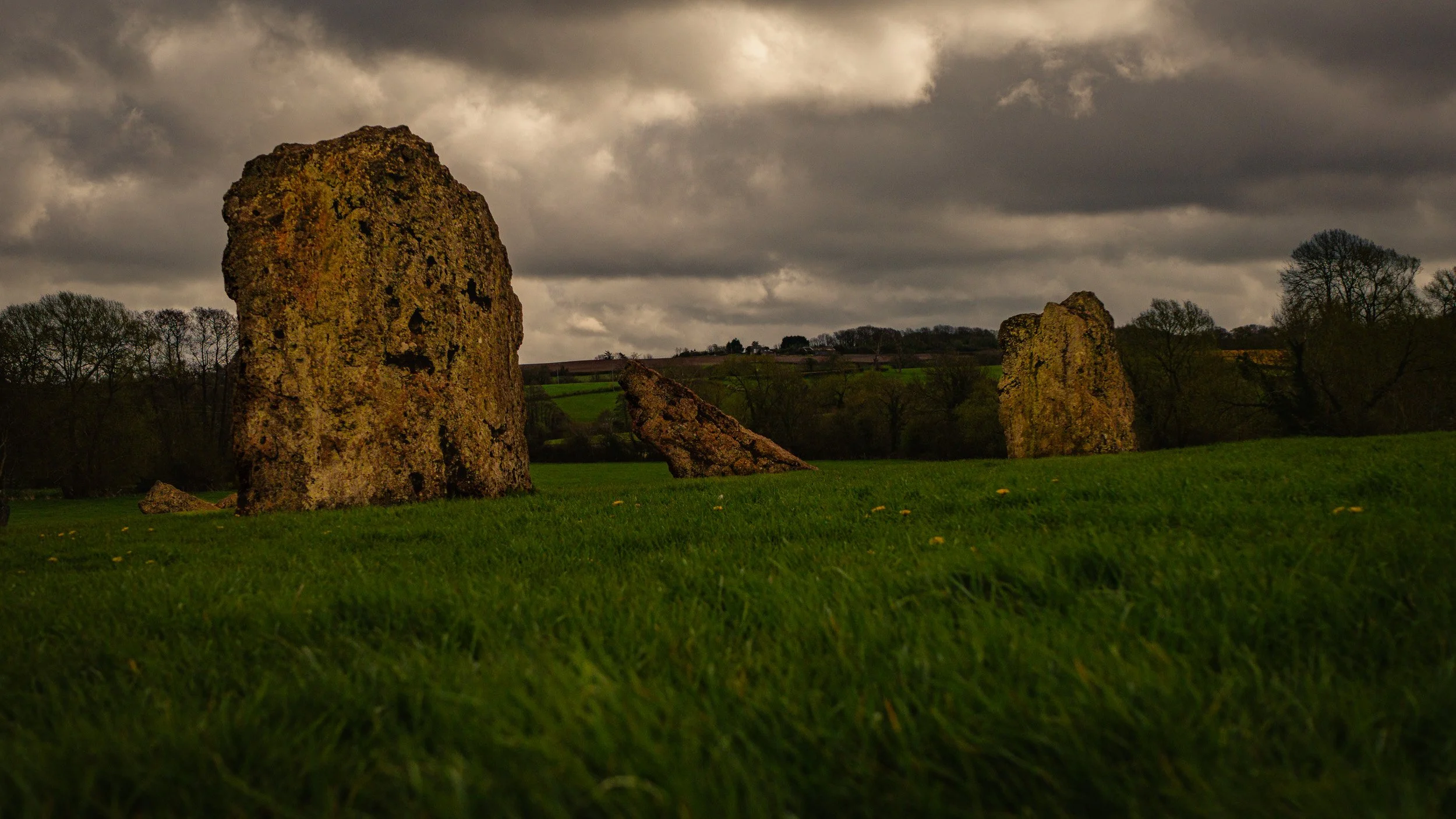 Large weathered standing stones in a grassy field with a few smaller stones, trees, hills, and dark cloudy sky in the background.