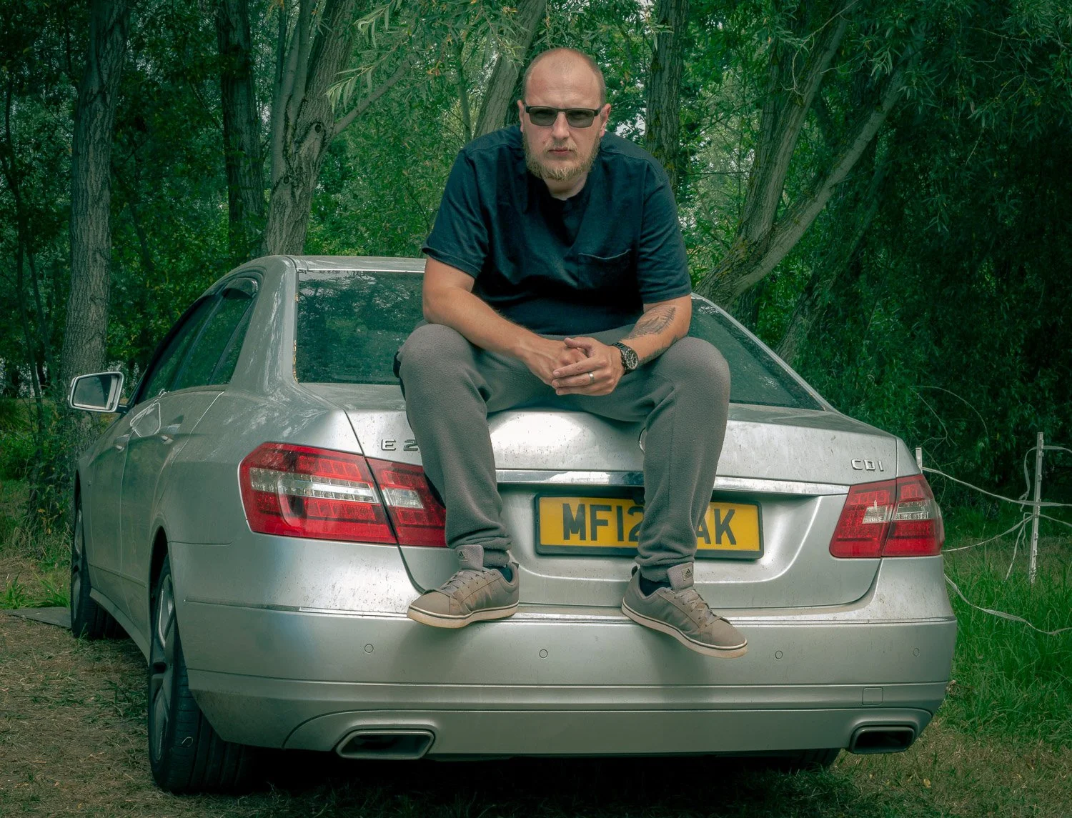 A man with sunglasses sitting on the trunk of a silver Mercedes-Benz E-Class car in a wooded area.