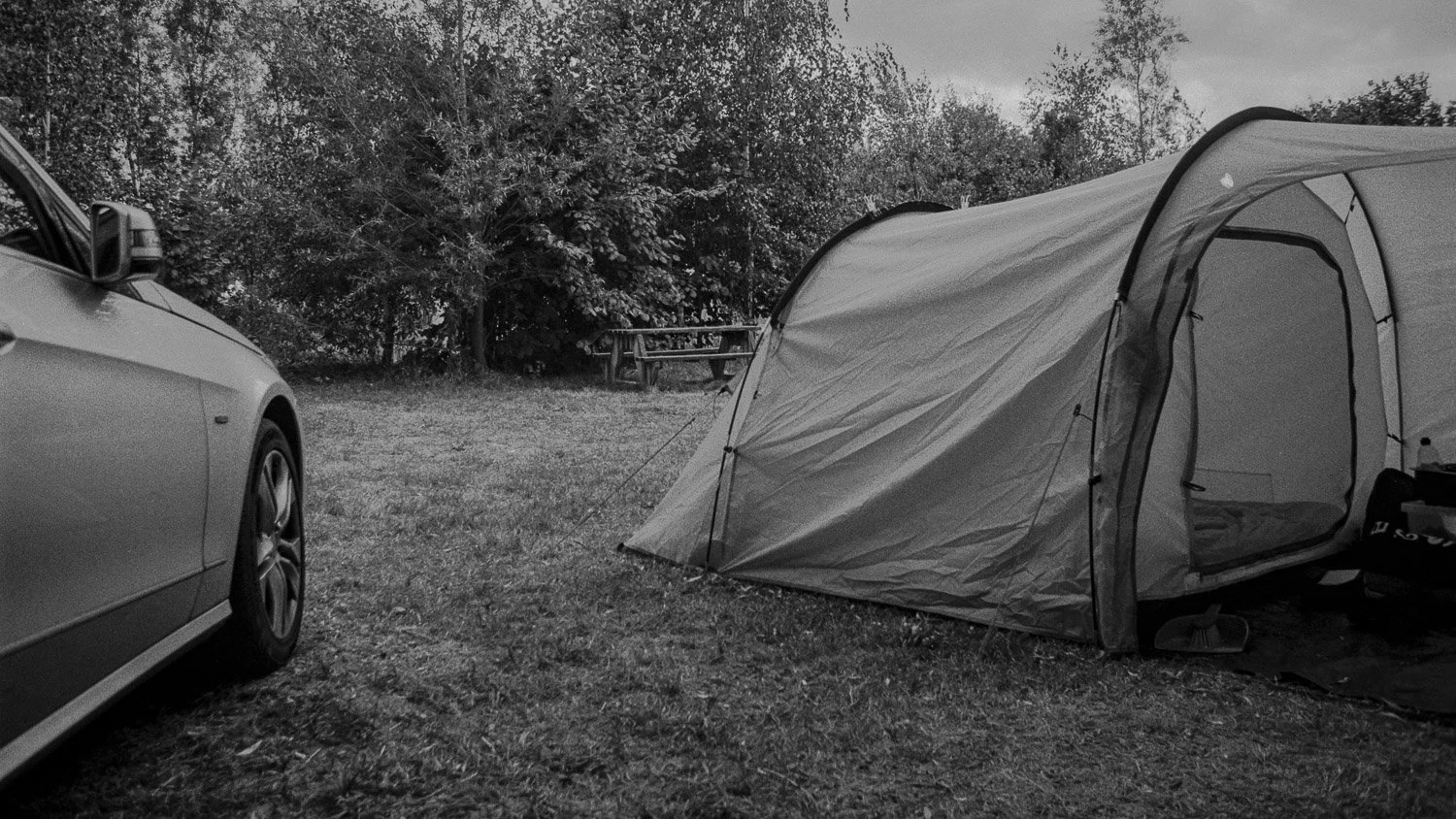 A camping scene with a car on the left and a tent on the right in a grassy area, with trees in the background.
