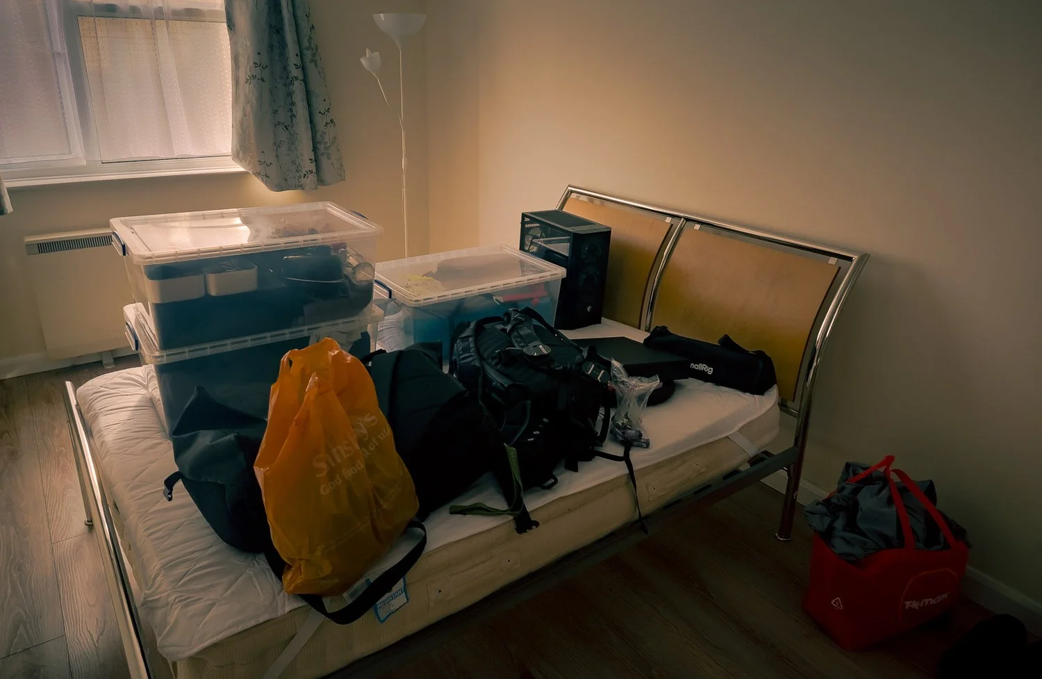 A hospital bed covered with various bags, backpacks, and storage containers, with additional boxes and equipment on site, next to a window with floral curtains and a standing lamp.