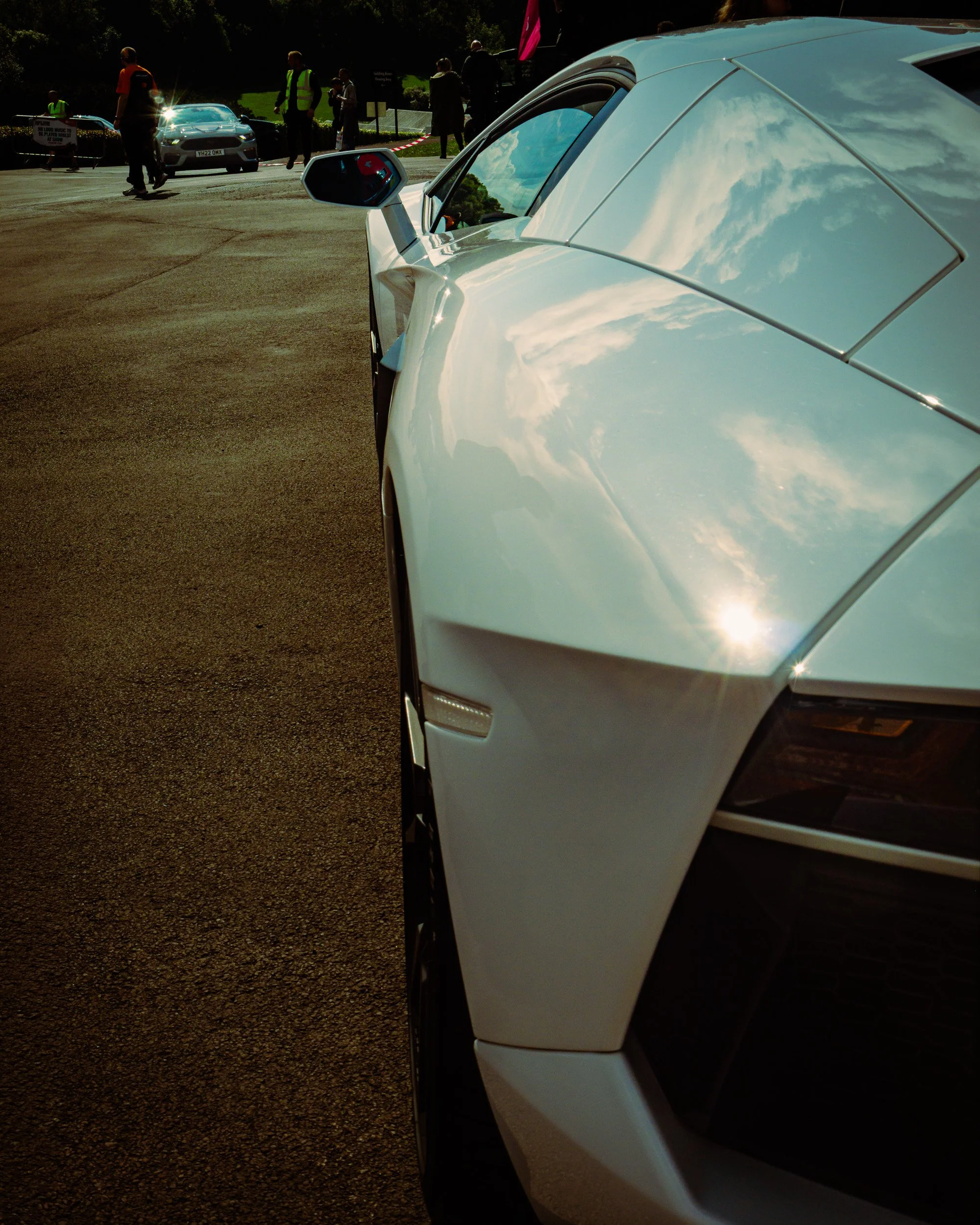 Close-up of the front side of a white sports car parked outdoors with reflections of clouds on its surface. In the background, there are people standing near other cars, some wearing reflective vests, and a sign indicating a holding area.
