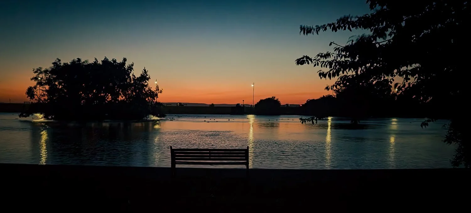 Bench facing a calm river during sunset or sunrise, with silhouetted trees and streetlights reflecting on the water.