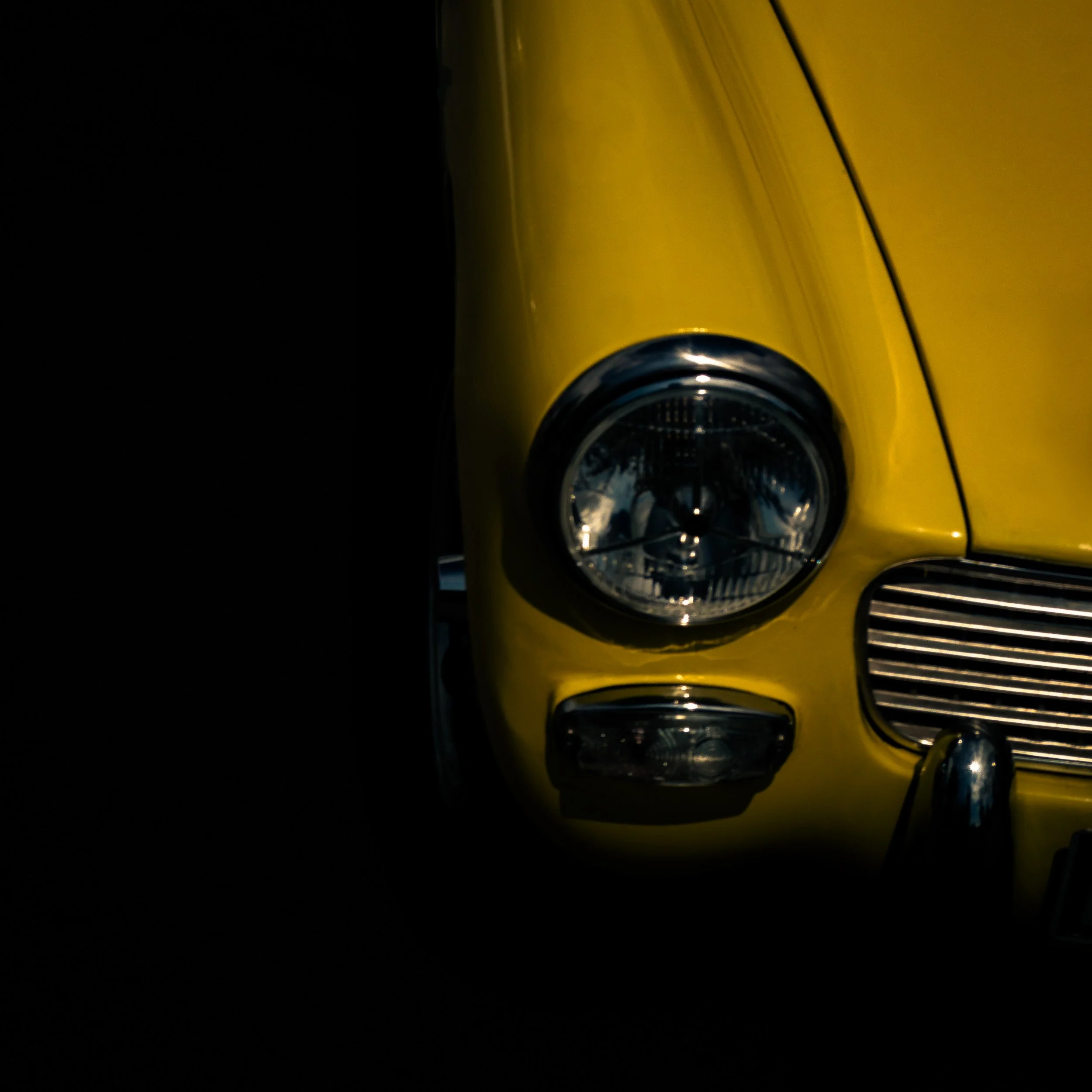Close-up of the front part of a yellow car, focusing on the headlight and grille, set against a dark background.