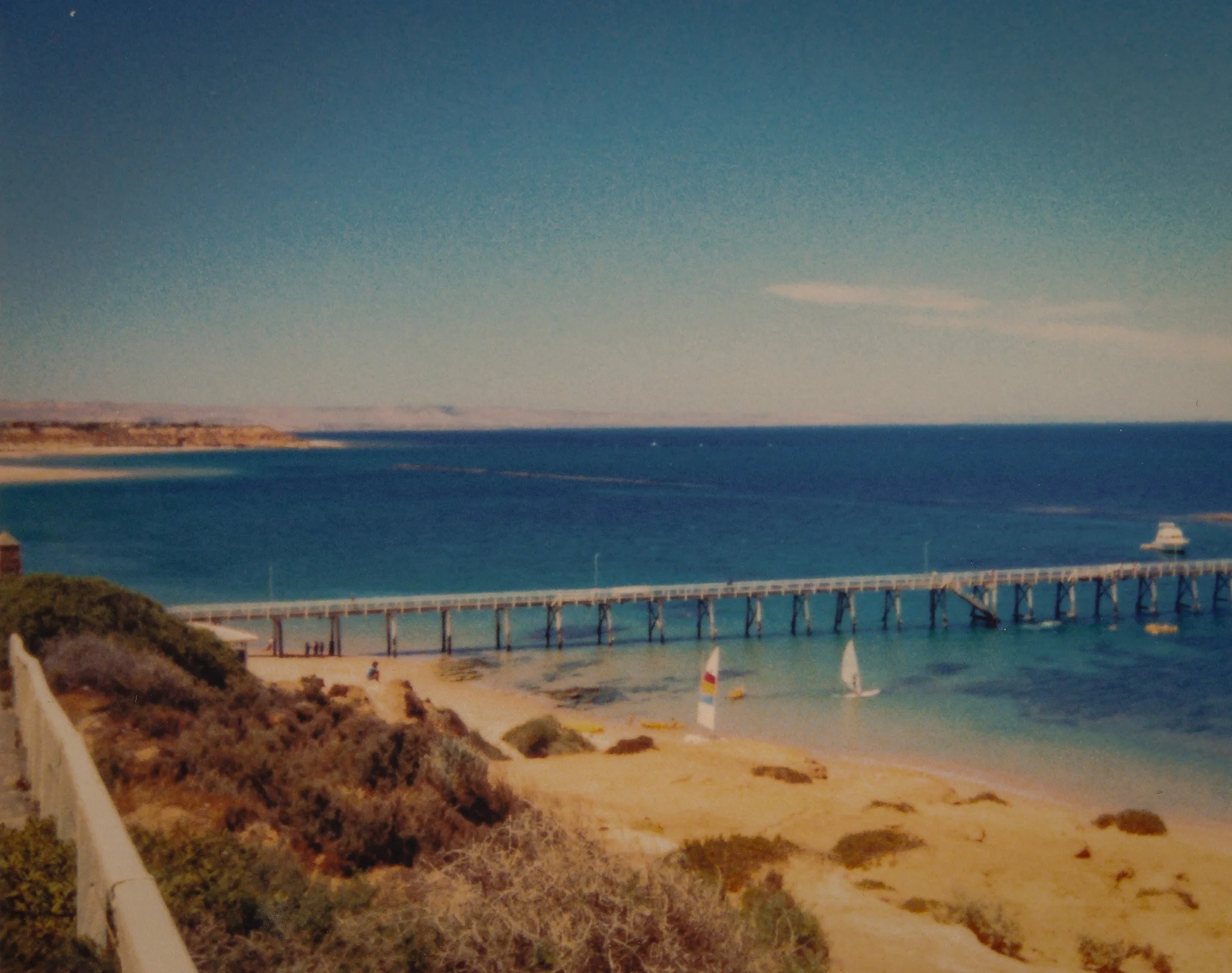A beach scene with sailboats and yachts, a long bridge over the ocean, and a distant landmass under a clear sky.