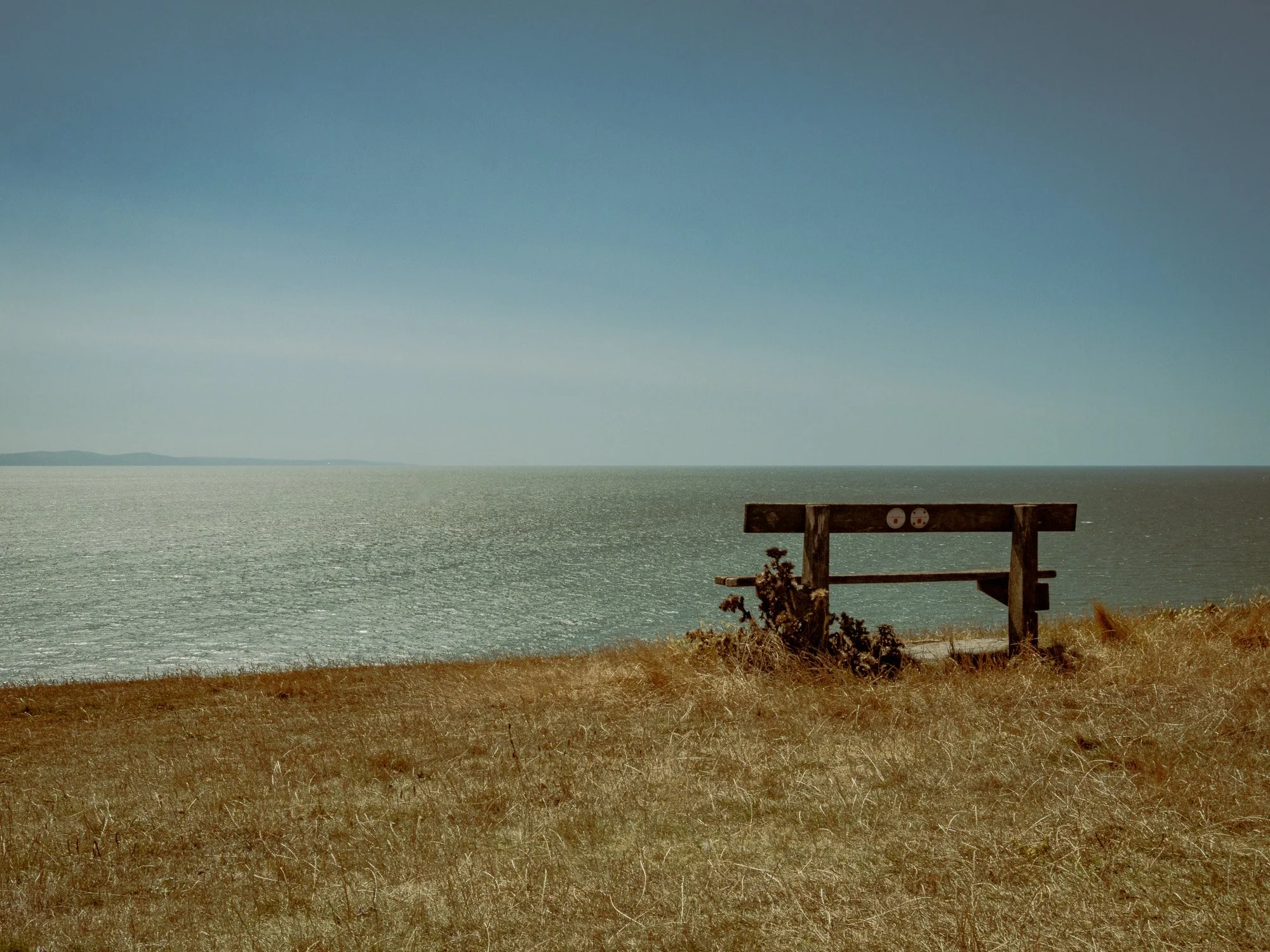 A bench overlooking a large body of water during daytime with a clear sky, some clouds, and distant land on the horizon.
