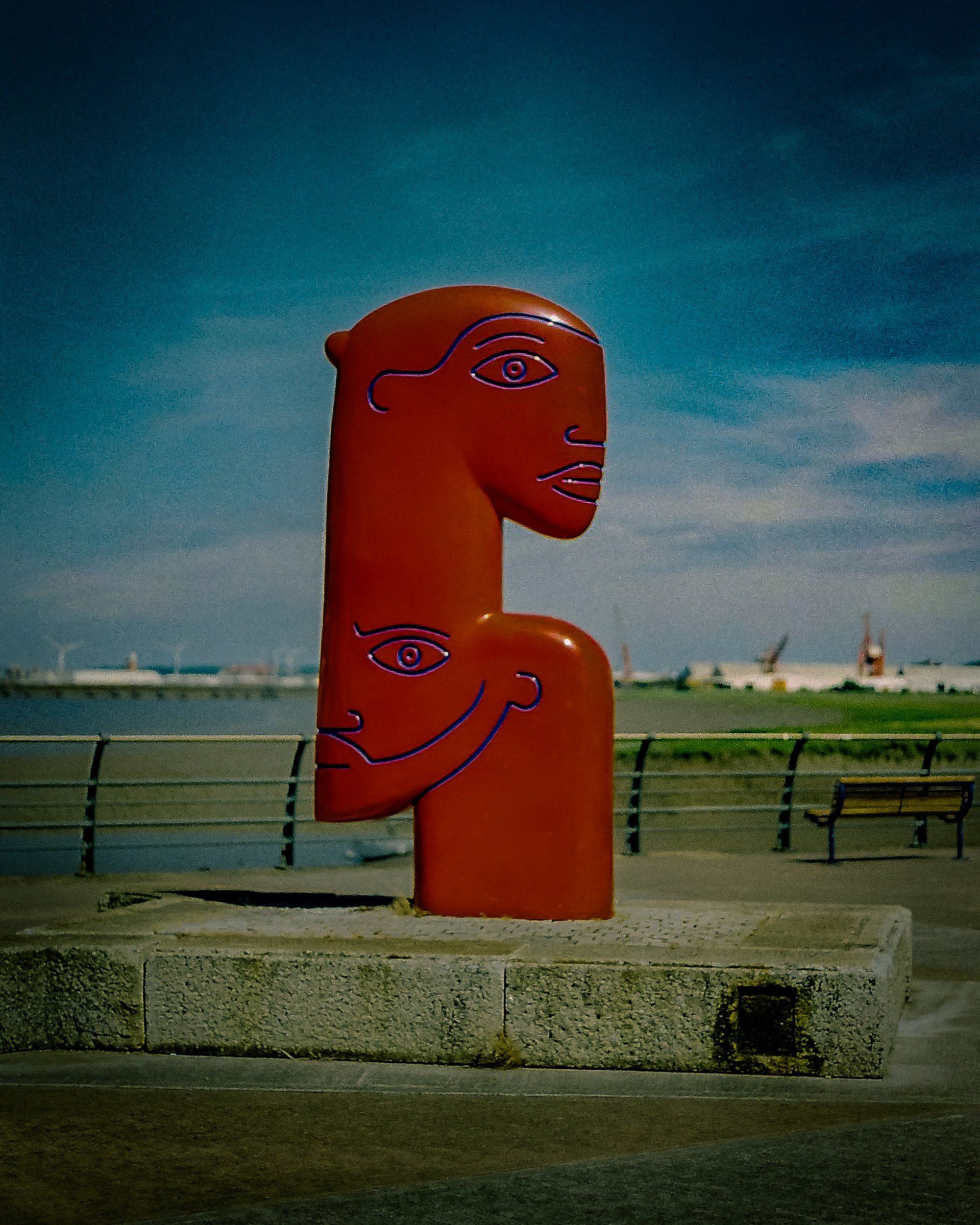 A tall orange sculpture of a human head with neon outline drawings of eyes, noses, mouths, and other facial features, positioned outdoors near a waterfront with cranes and buildings in the background, under a dark sky.