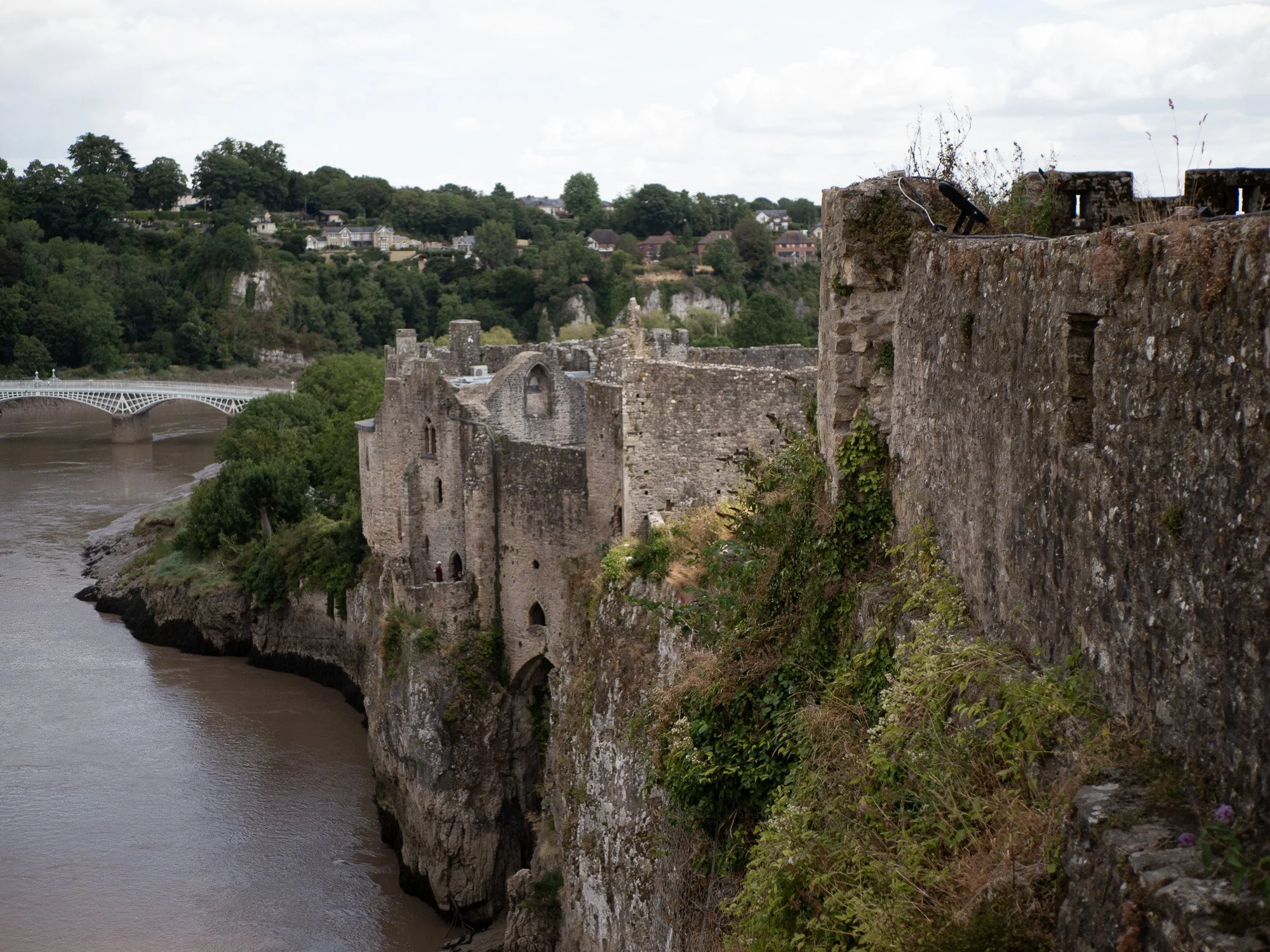 Ancient stone castle ruins on a cliff overlooking a river with a small town and a distinctive white arch bridge in the background, under a partly cloudy sky.