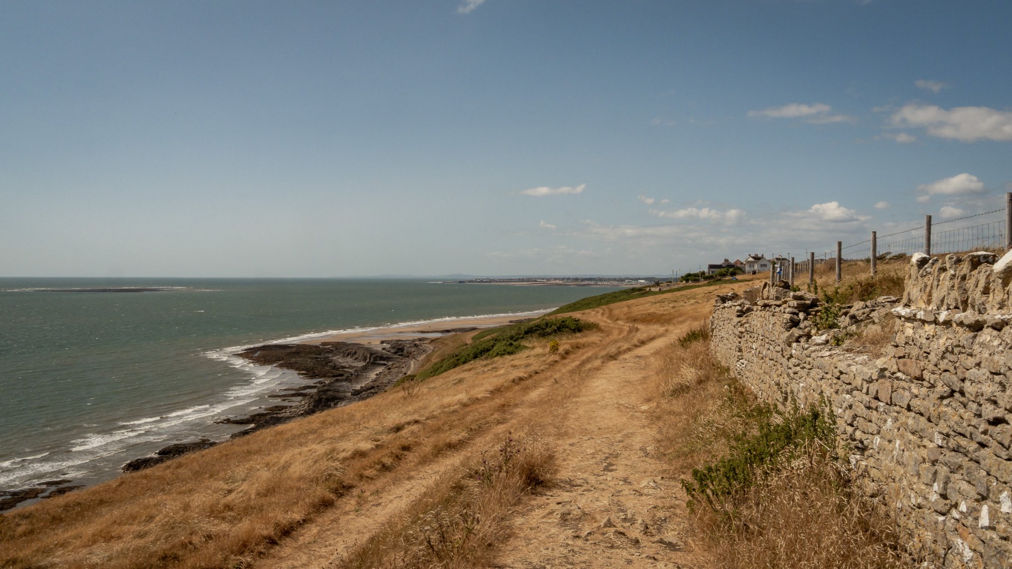 A coastal view featuring a dirt trail along a stone wall on the right, leading to homes in the distance, overlooking a rocky shoreline and ocean under a partly cloudy sky.