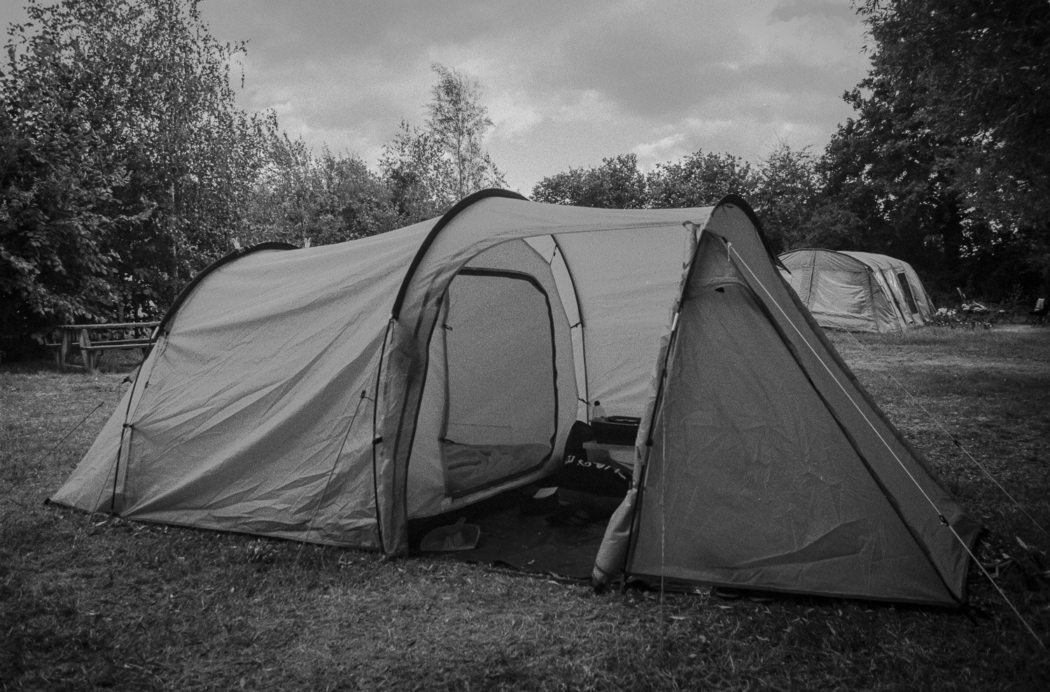 A black-and-white photo of a camping scene with a large tent in the foreground and smaller tents in the background, set on a grassy area surrounded by trees.