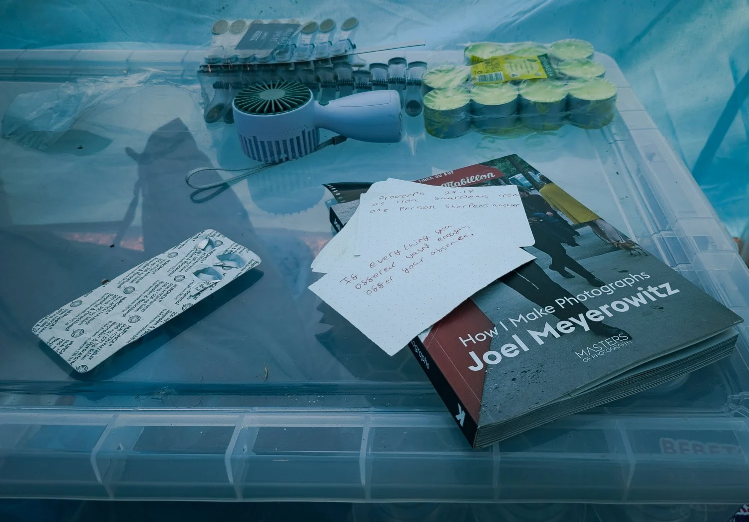 A plastic storage box containing a book titled "How I Make Photographs" by Joel Meyerowitz, notes, a small fan, a sheet of paper with handwritten text, a package of yellow sponges, and a pack of batteries.