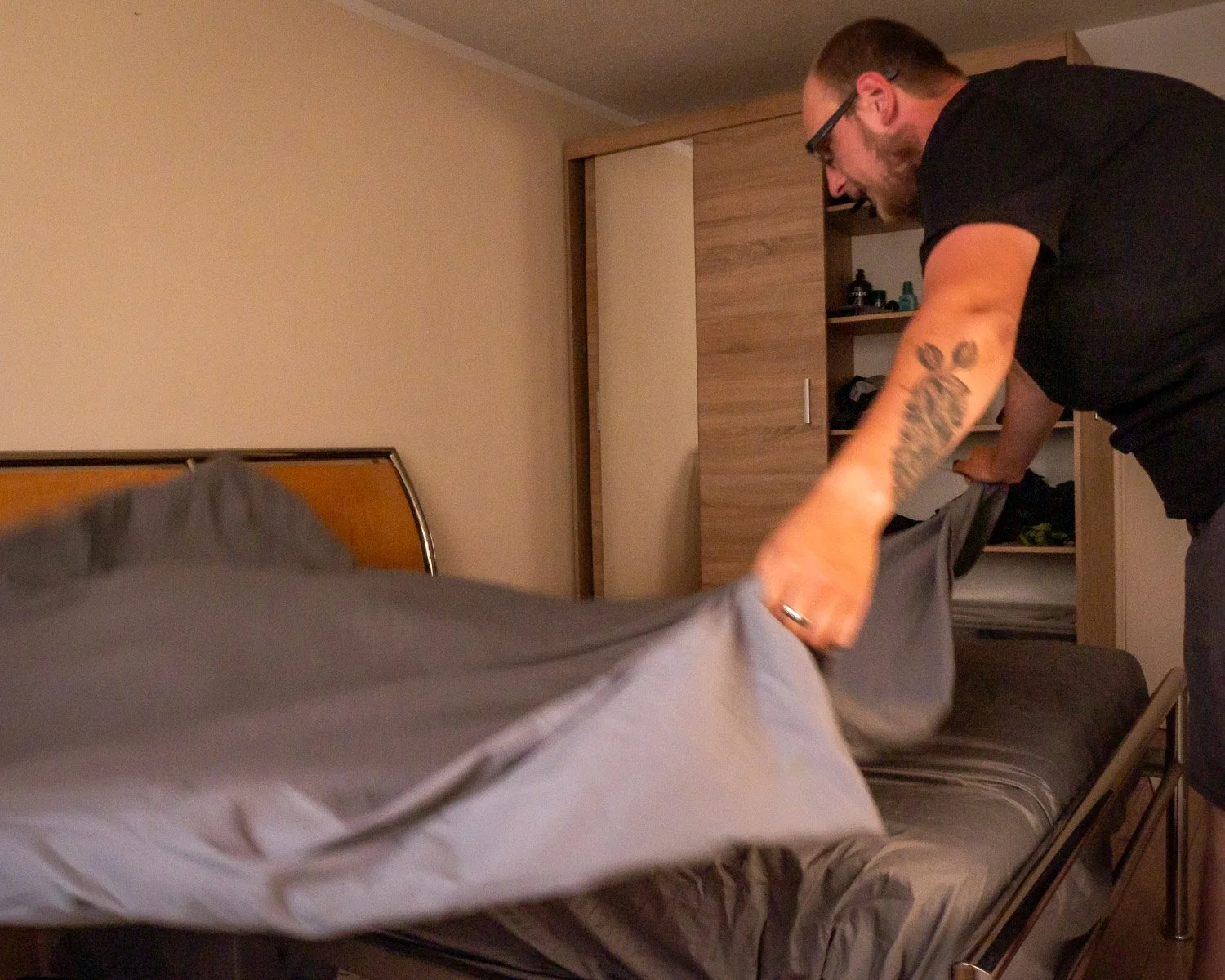 A man with glasses and tattoos is making a bed with gray sheets in a bedroom with beige walls and a wooden wardrobe.