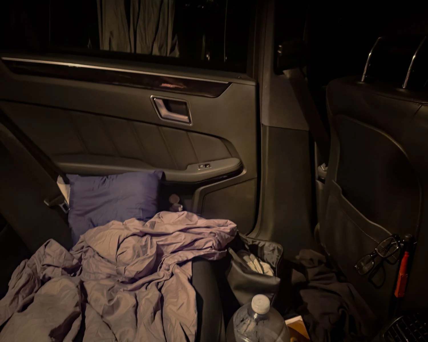 A car interior at night with the back seat and door visible, cluttered with clothes, a water bottle, glasses, and other items.