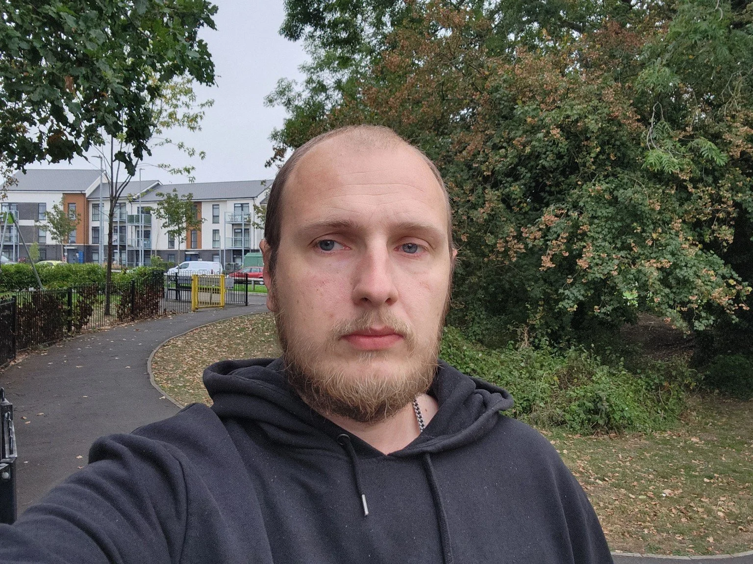 A man with a beard and long hair taking a selfie in a park with trees and modern residential buildings in the background.