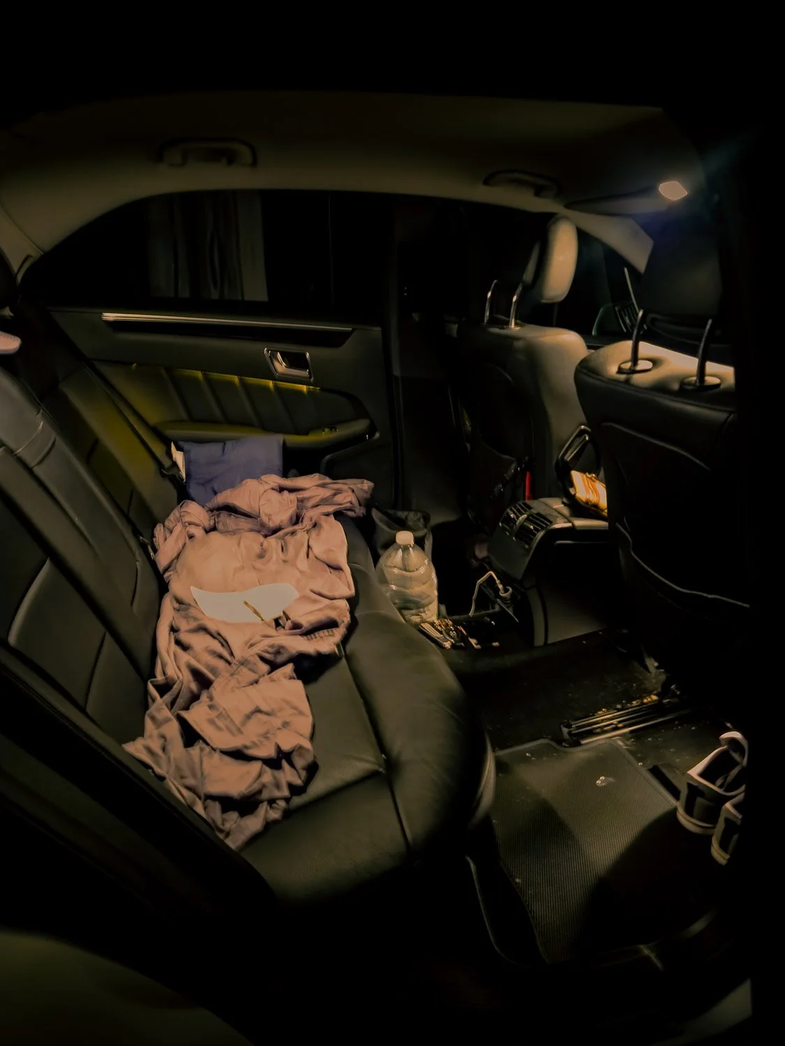 The back seat of a car with scattered clothes, a pillow, a water bottle, and some paper on it, with some items and maps visible in the front.