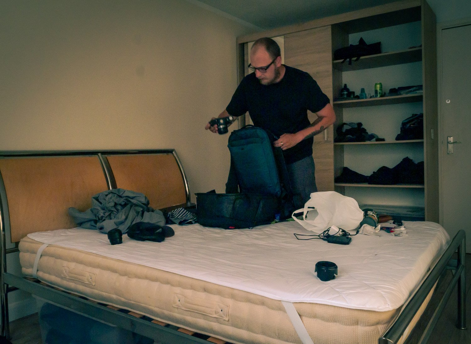 Man packing or unpacking belongings on a bed in a bedroom with shelves and an empty mattress.