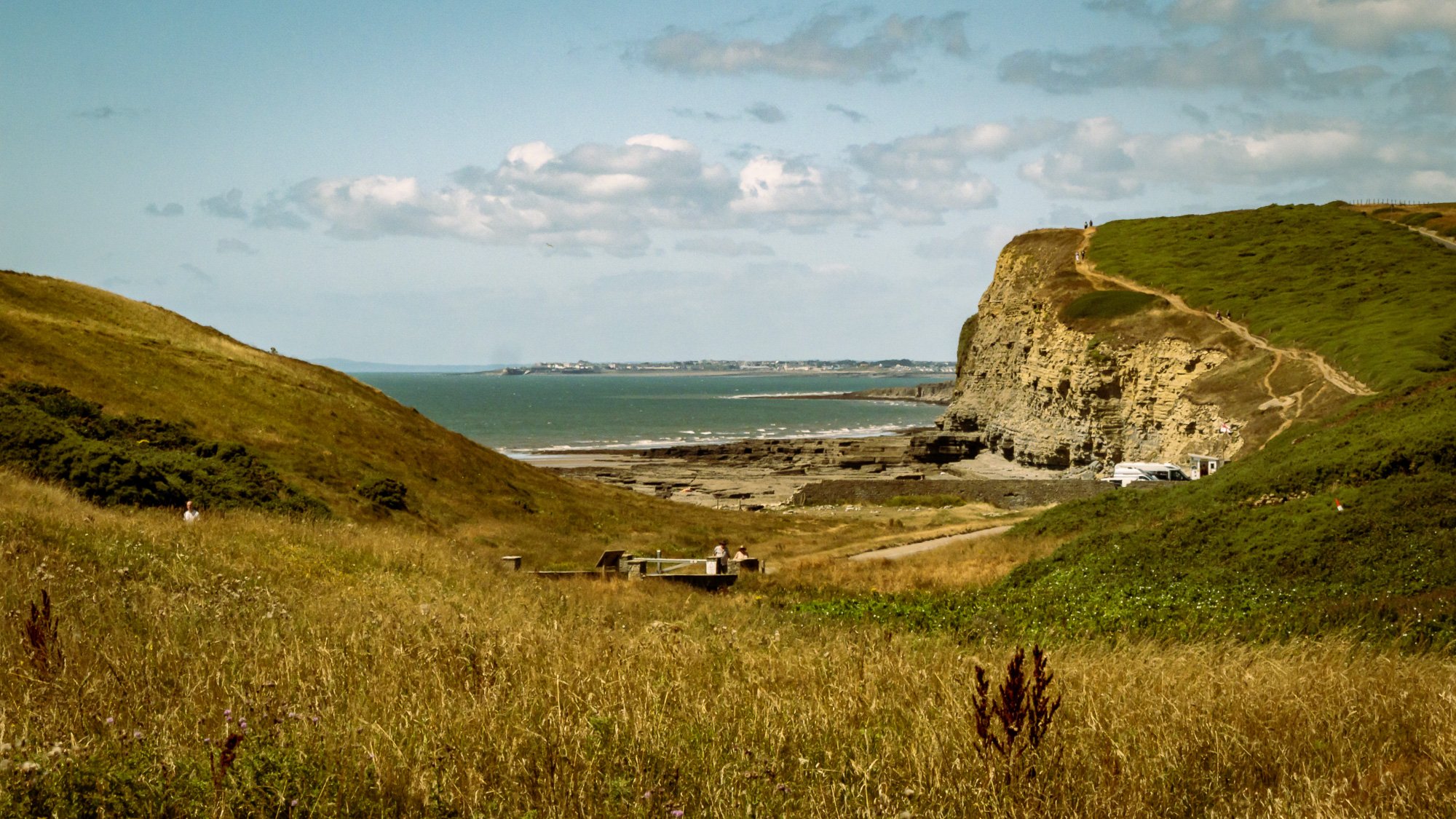 A coastal landscape with green grassy hills, a sandy beach, and cliffs facing the ocean, under a partly cloudy sky, with a few people walking along the trails on the hills.