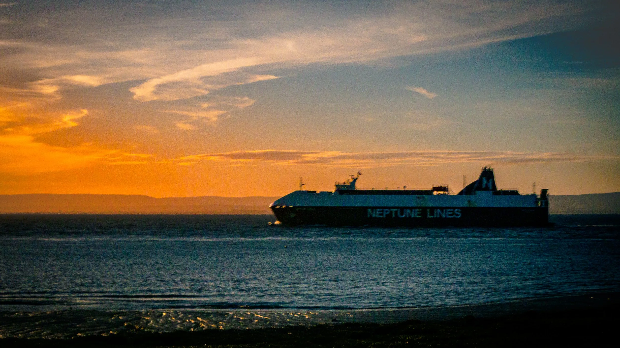 A large ferry boat with 'Neptune Lines' written on its side sailing on calm water during sunset with clouds in the sky.