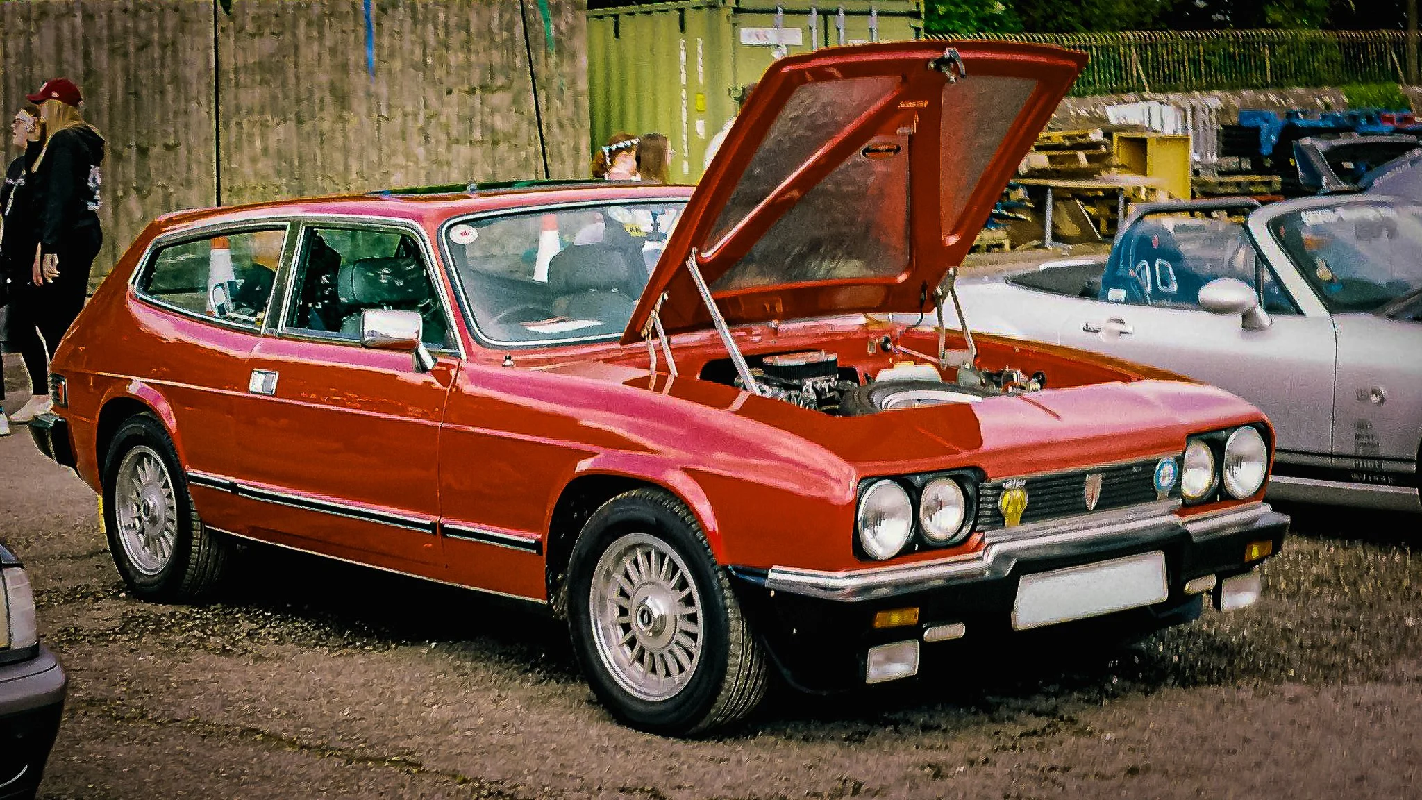 A red vintage car with an open hood parked among other vehicles at a car show, with several people in the background.
