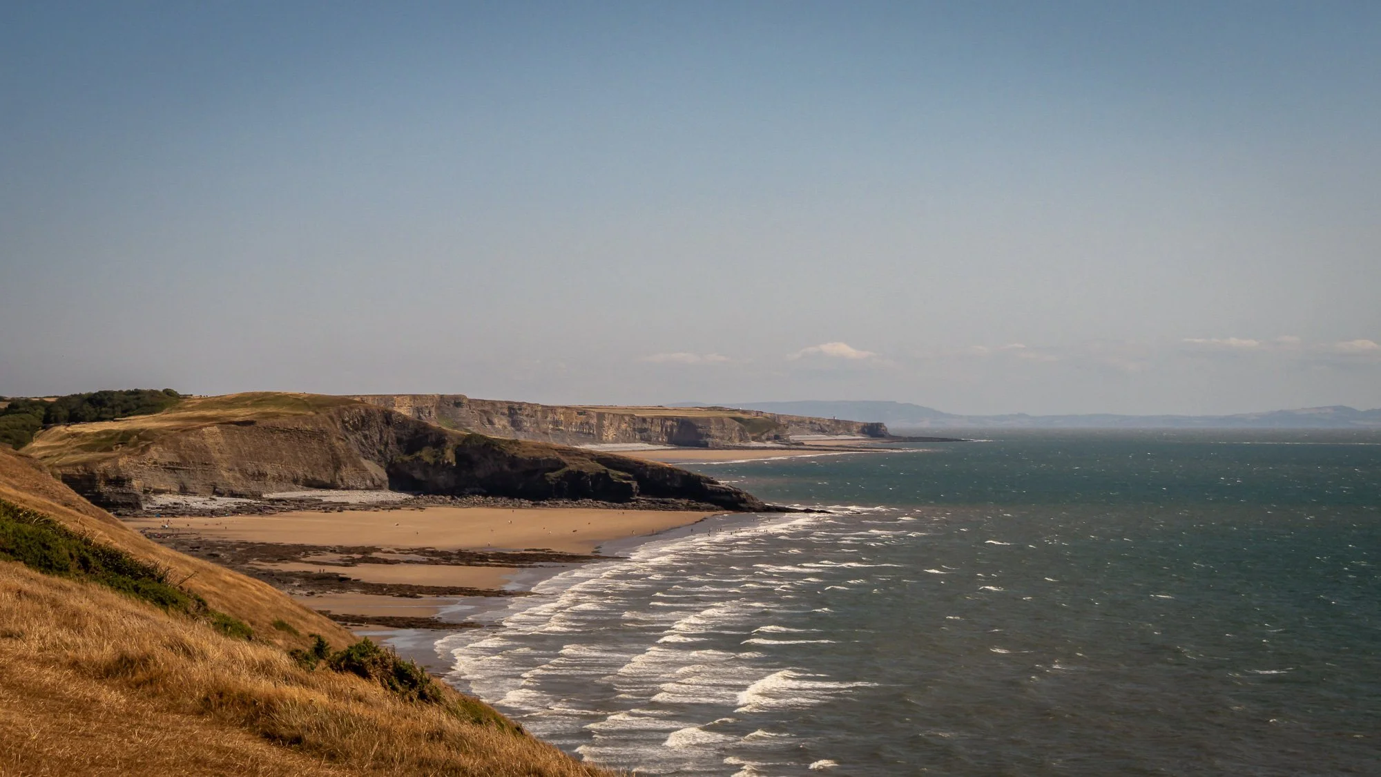 Cliffs along a sandy beach with waves, under a blue sky with a few clouds, seen from a grassy hillside.