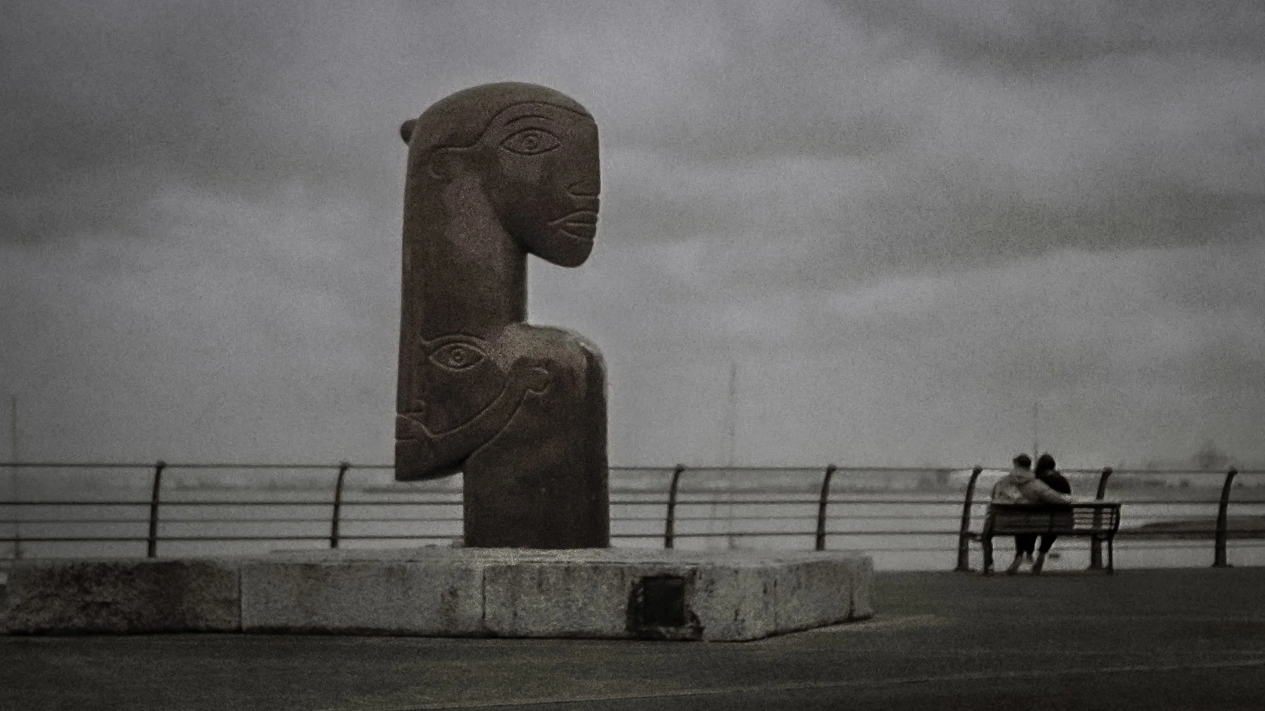 A large carved stone sculpture resembling a face with stylized eyes, nose, and mouth, stands on a platform outdoors. In the background, a fence and a cloudy sky are visible, with two people sitting on a bench.