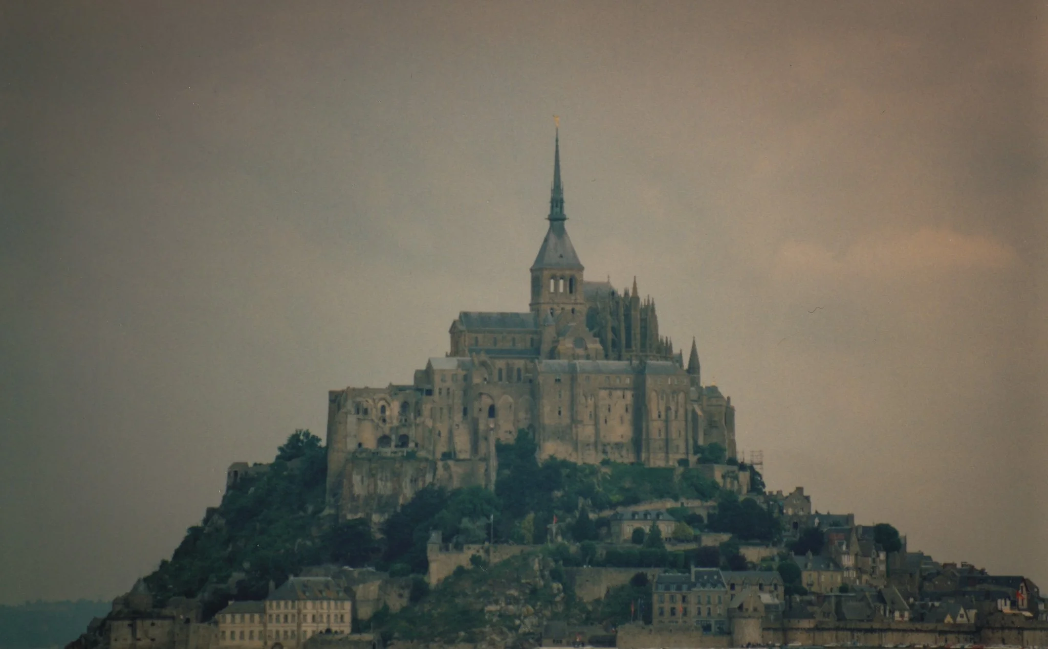 A historic castle built on a hilltop, overlooking a town beneath, with a tall spire and gothic architectural details, under a cloudy sky.