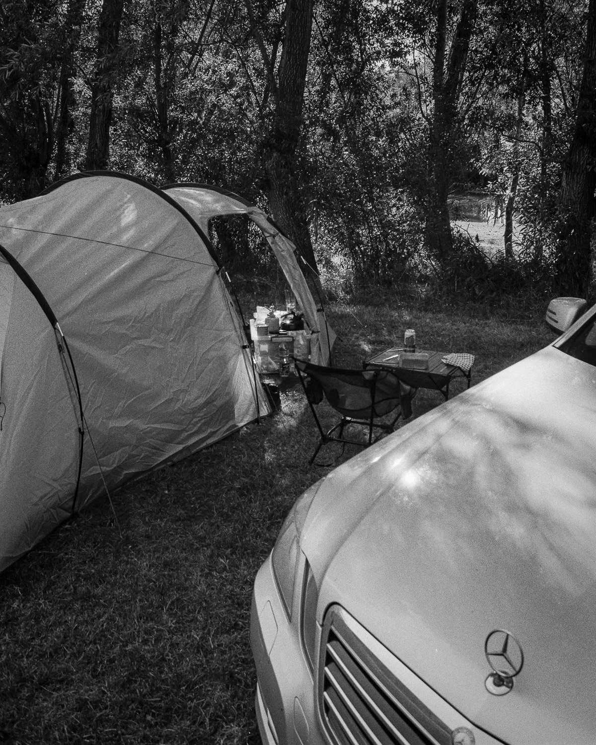A camping setup with two tents, a folding chair, a table with items, and a car in a wooded area.