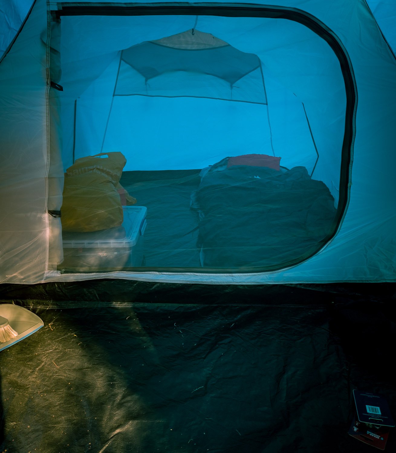 Interior of a camping tent with a bed, pillows, and clothing inside, viewed through a large clear window.