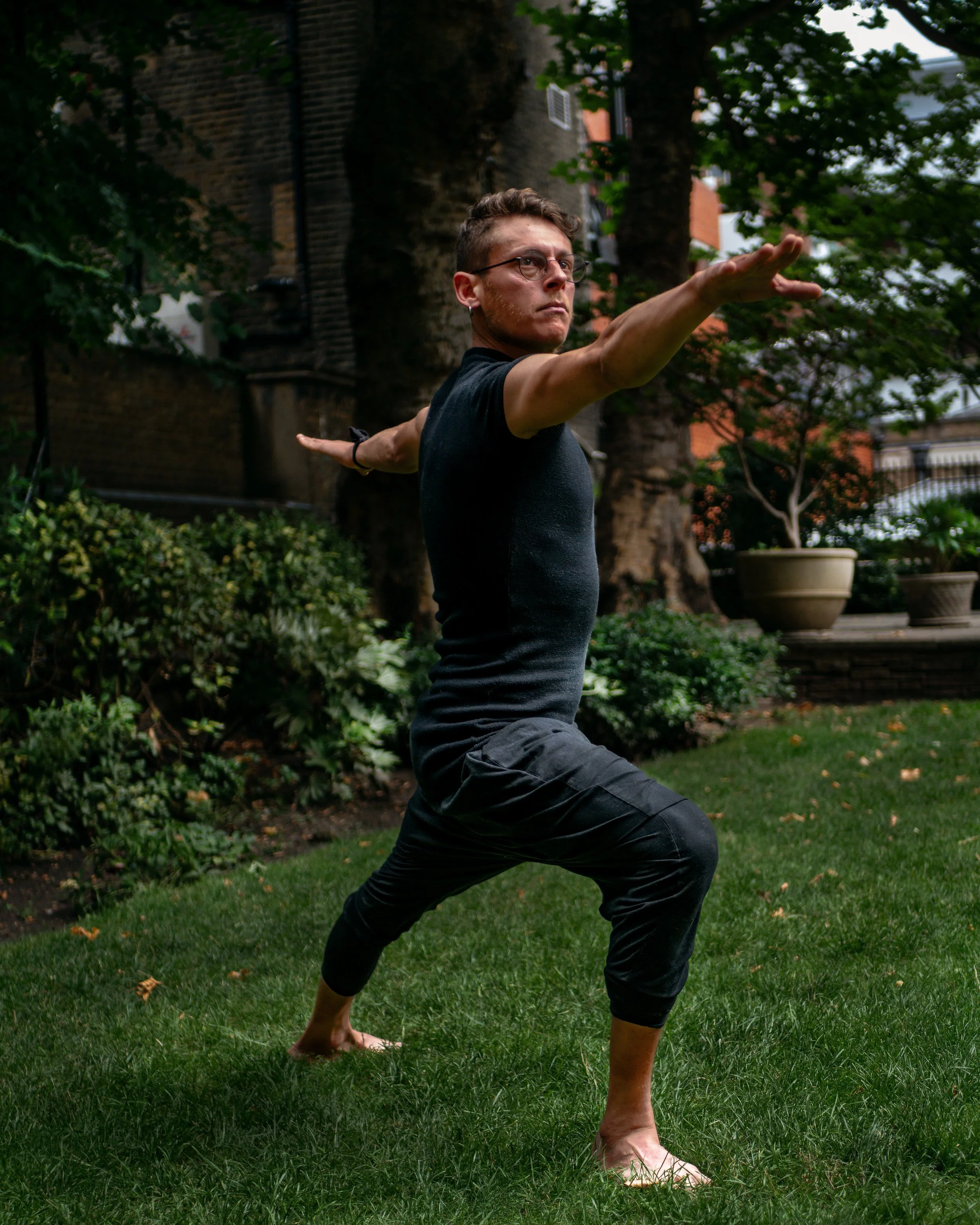 A young man practicing yoga outdoors in a garden, balancing in a warrior pose with arms extended and one knee bent, wearing glasses, a black shirt, and black pants, surrounded by green trees and plants.