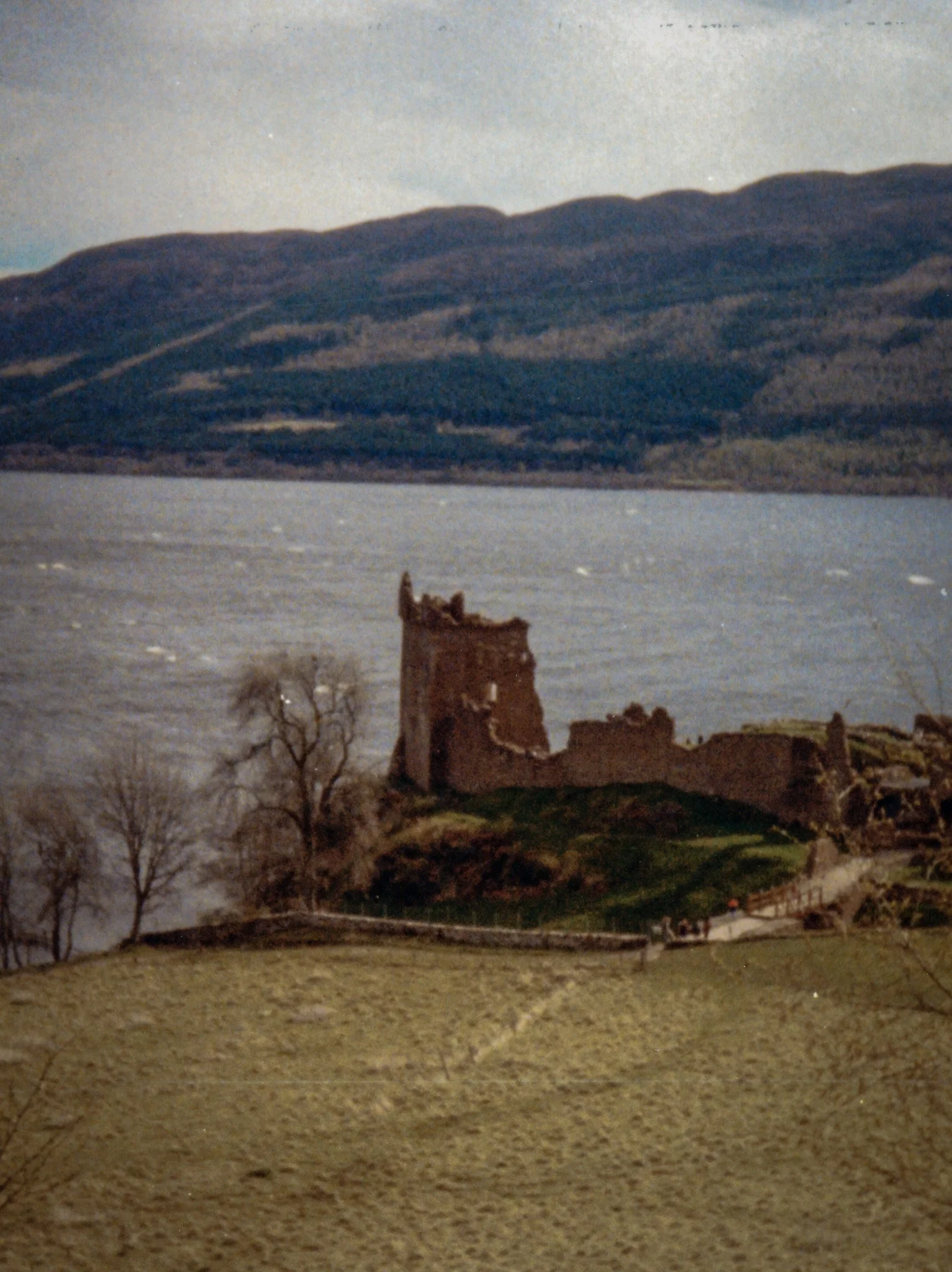 A landscape scene showing a body of water with a mountainous terrain in the background. In the foreground, there are remnants of a destroyed building with some leafless trees nearby.