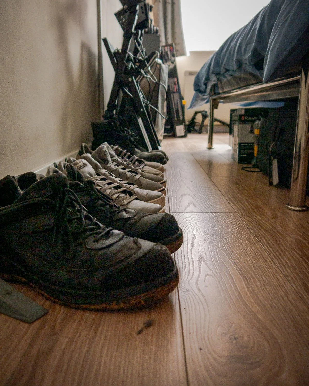 Multiple pairs of sneakers lined up on a wooden floor next to a wall, in a room with a bed, a desk, and various electronic equipment.